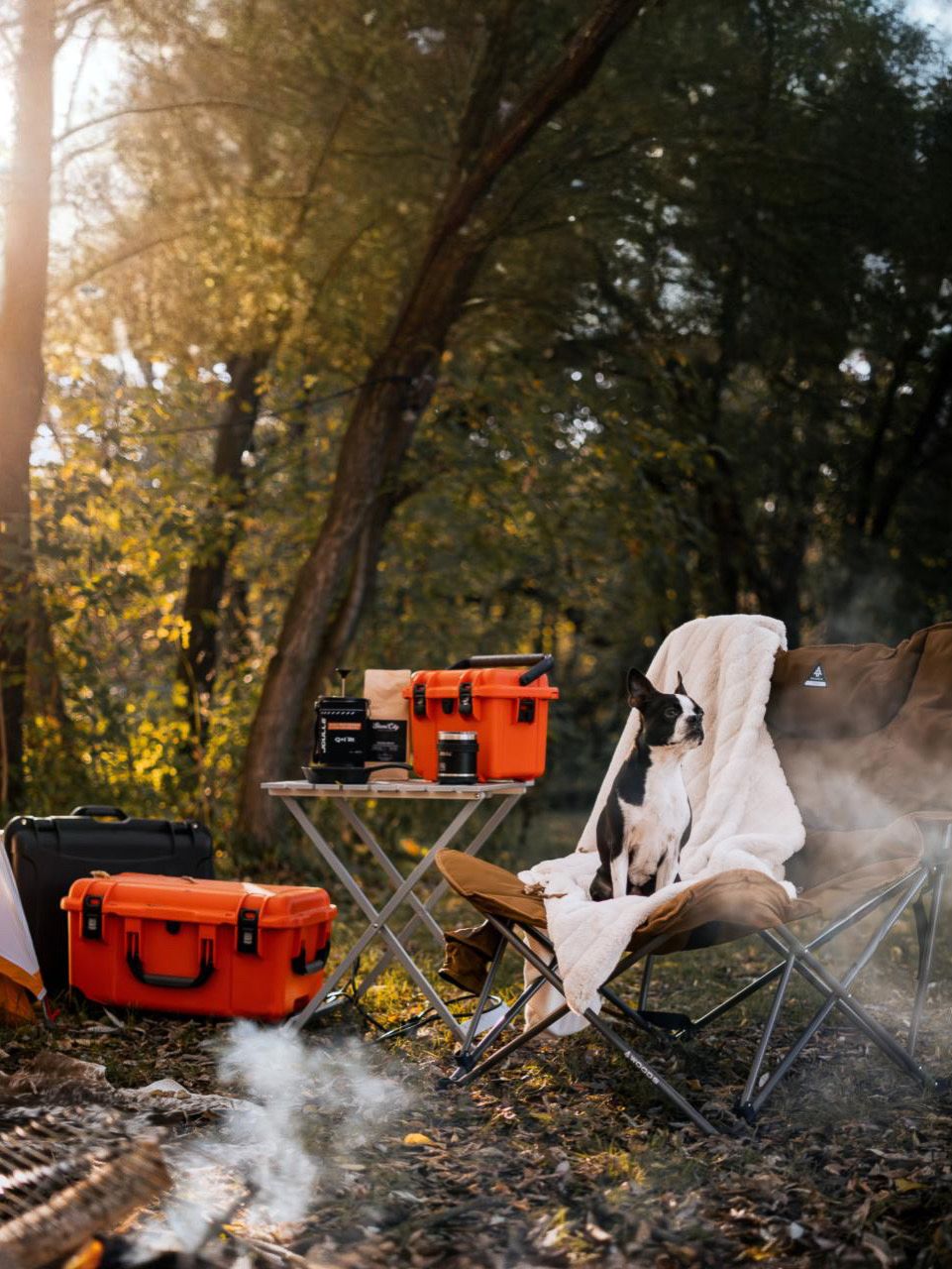 Smoke rises from camping setup with orange equipment case in autumn forest setting.