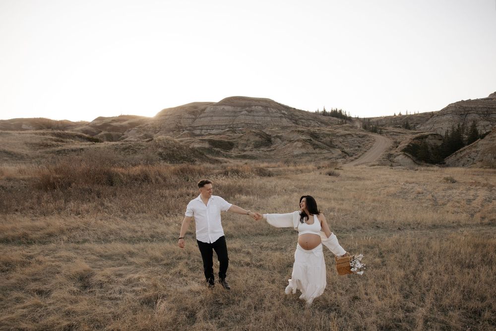 Pregnant couple holds hands walking through golden field with rolling hills at sunset during a maternity photo shoot in calgary