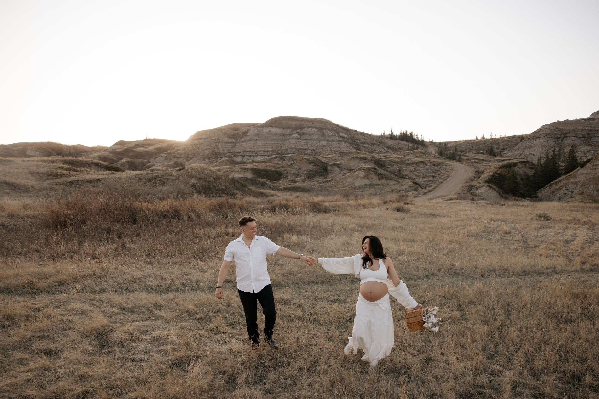 Pregnant couple holds hands walking through golden field with rolling hills at sunset during a maternity photo shoot in calgary