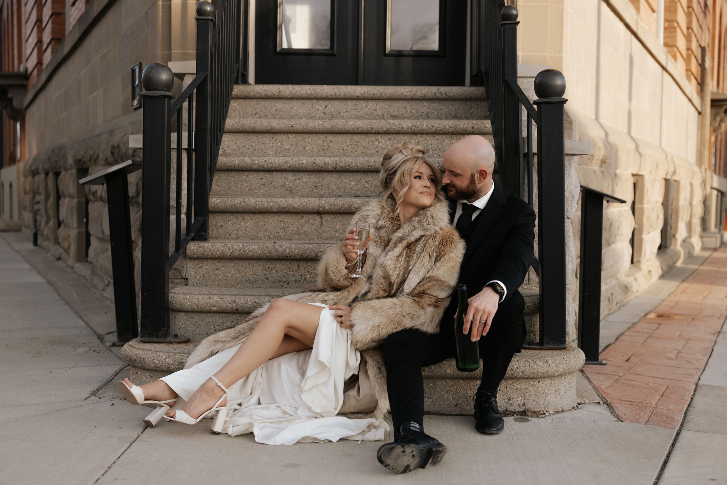 Bride and groom sitting on city steps, drinking champagne after eloping in alberta