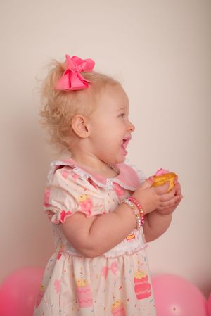 Toddler in pink bow and dress laughs while holding a treat at a birthday celebration.