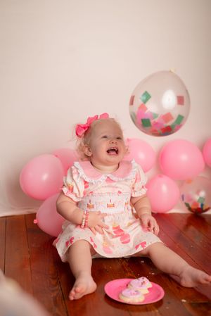Happy toddler in pink dress celebrates with balloons and cake at birthday party.