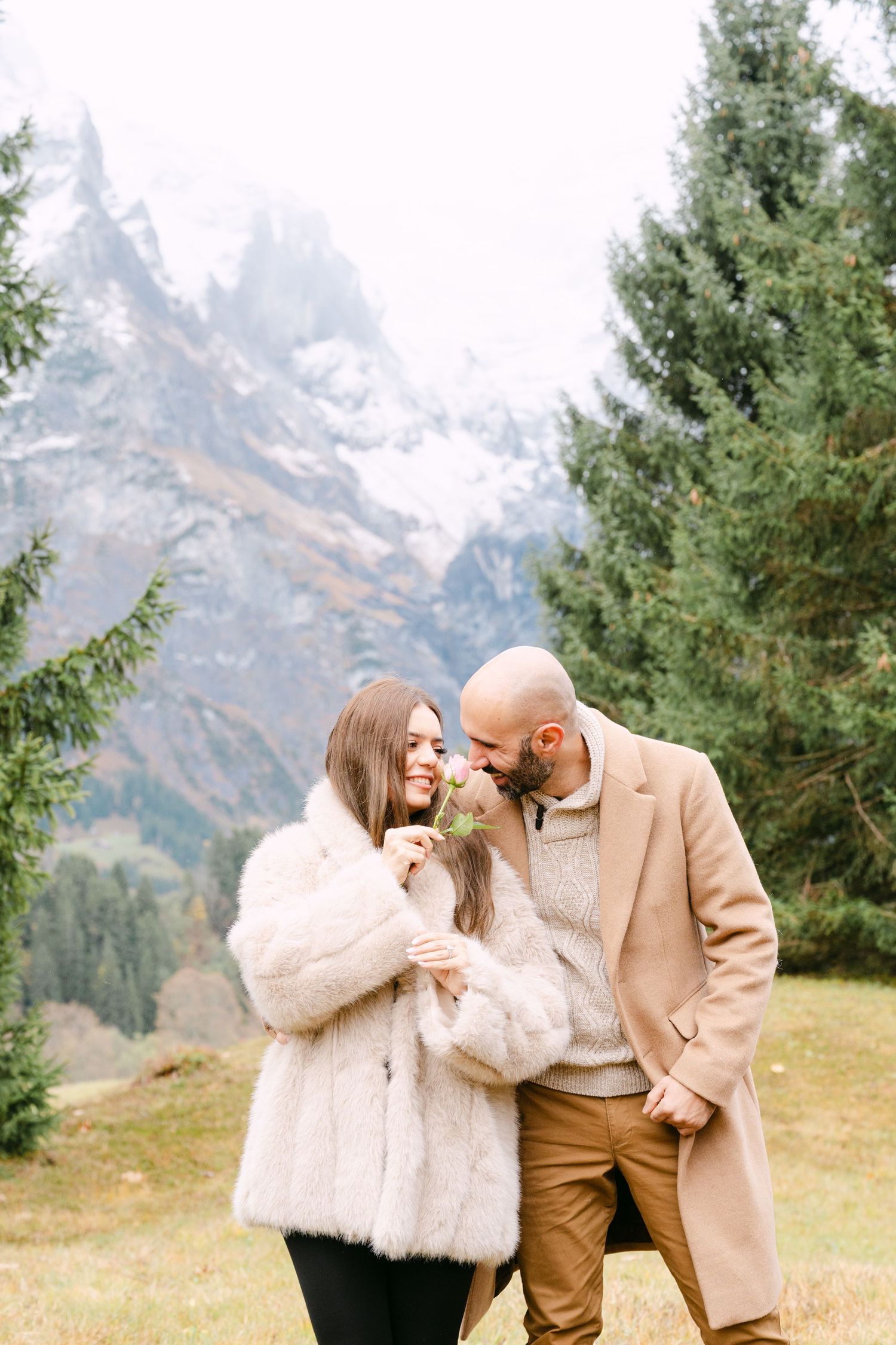 A couple shares tender moments in the mountains with evergreen trees and snow-capped peaks in the background.