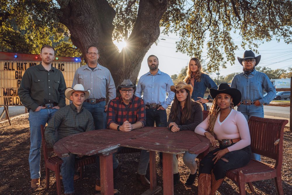 A group of people in western attire gathered around a wooden table outdoors under a tree during sunset.
