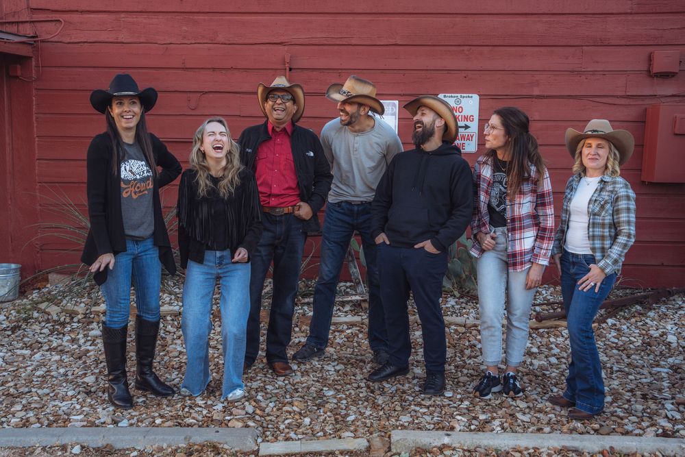 A group of people wearing western attire and cowboy boots stand against a red barn wall in autumn.