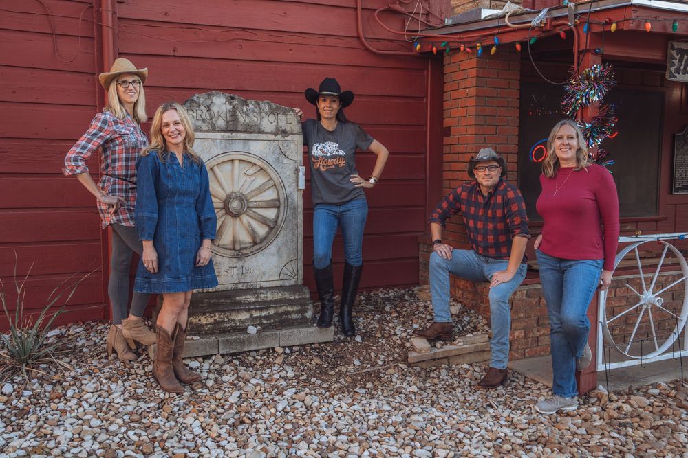 A group of people pose in front of a rustic red barn with decorative wagon wheels and vintage farm equipment.