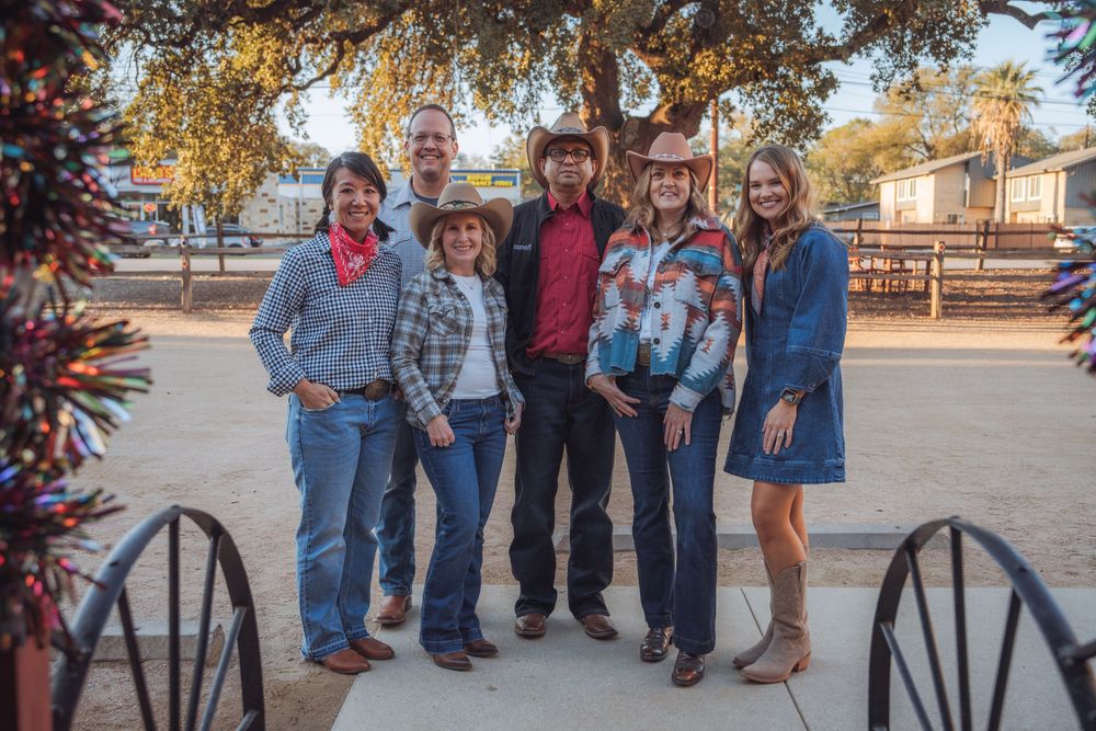 A group of people in casual attire pose together outdoors near an antique wagon wheel.