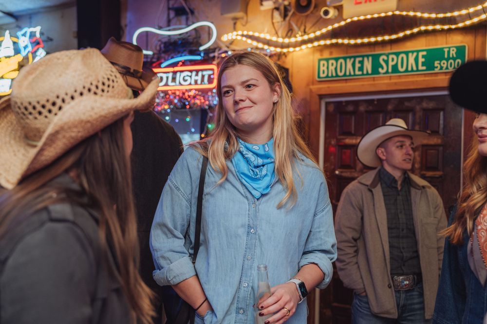 People socializing at a western-themed bar with neon signs and cowboy attire in the background.
