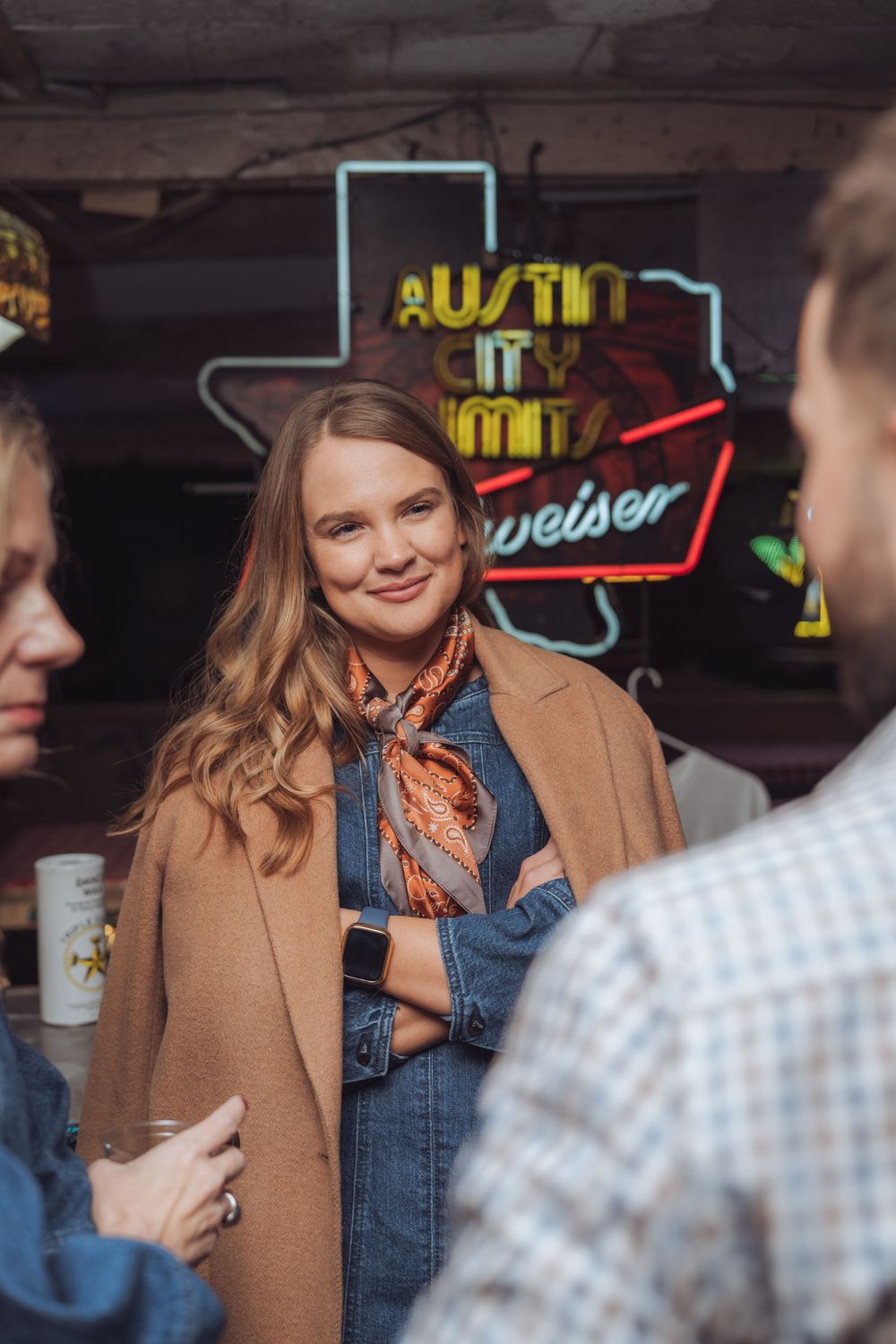 A casual gathering at an Austin bar with neon signs, featuring someone in a camel coat and denim jacket.