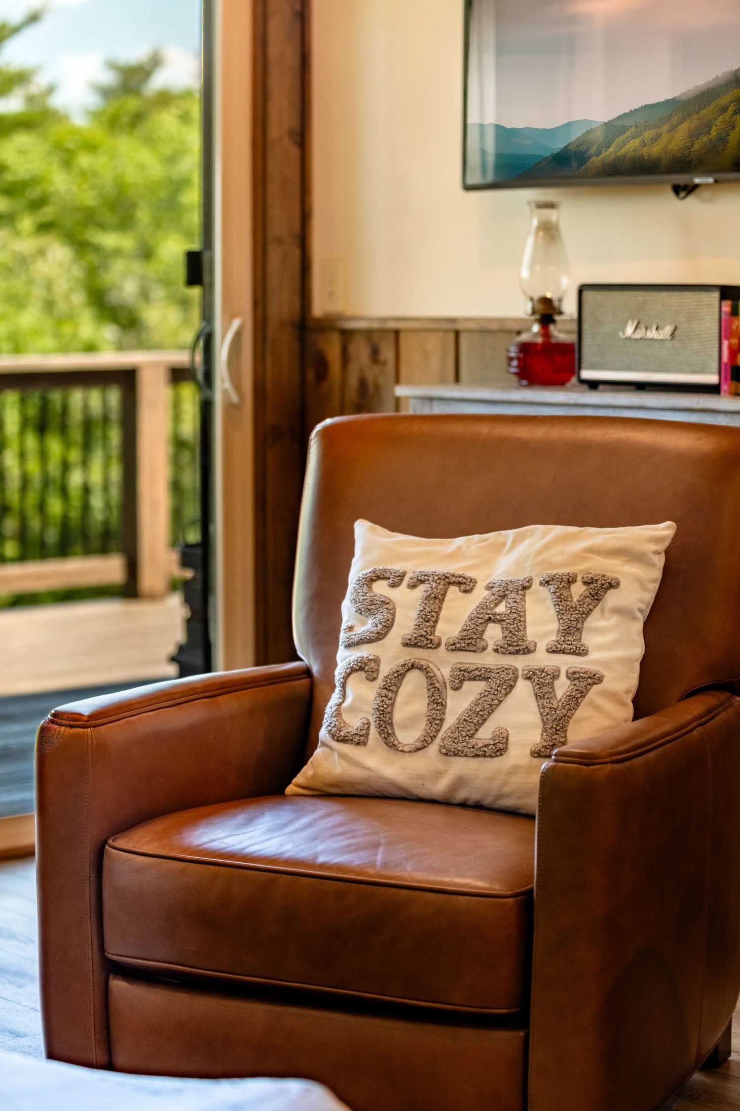 Leather armchair with a pillow that says stay cozy and a patio door leading to an outdoor deck overlooking trees.