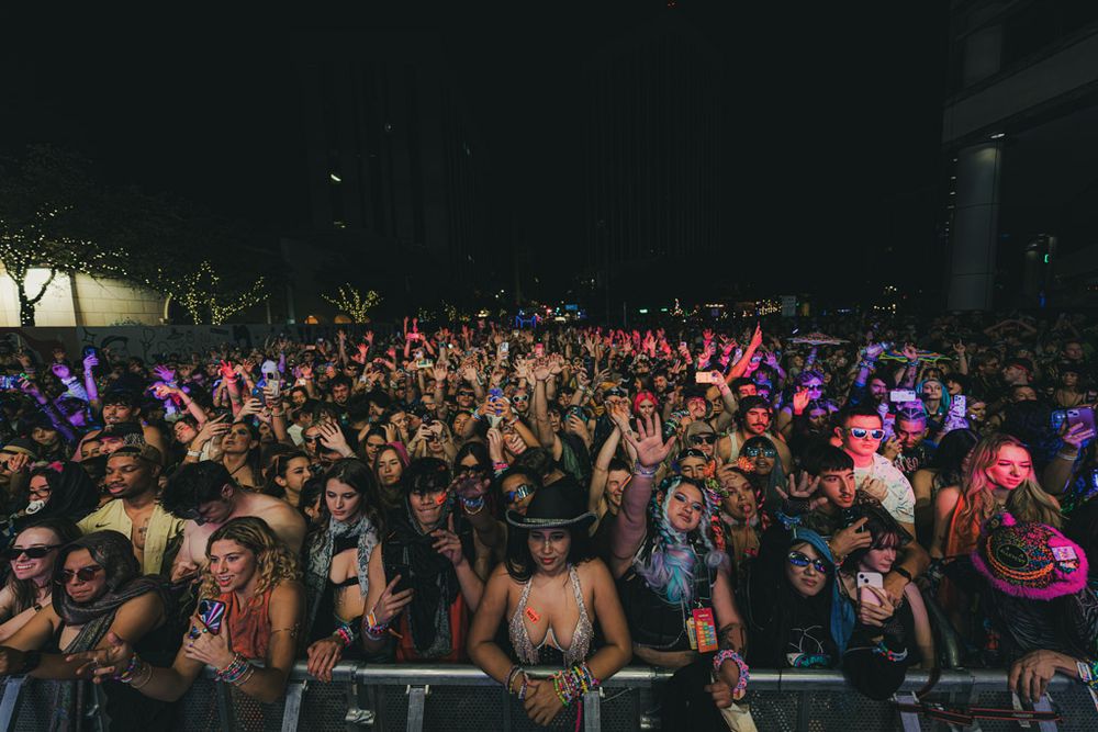 Large crowd of concert attendees with raised hands enjoying a nighttime music festival under colorful lights.