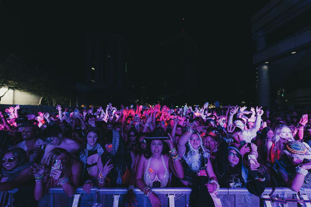 A large crowd of concert attendees stands in a dark venue illuminated by purple and blue stage lighting.