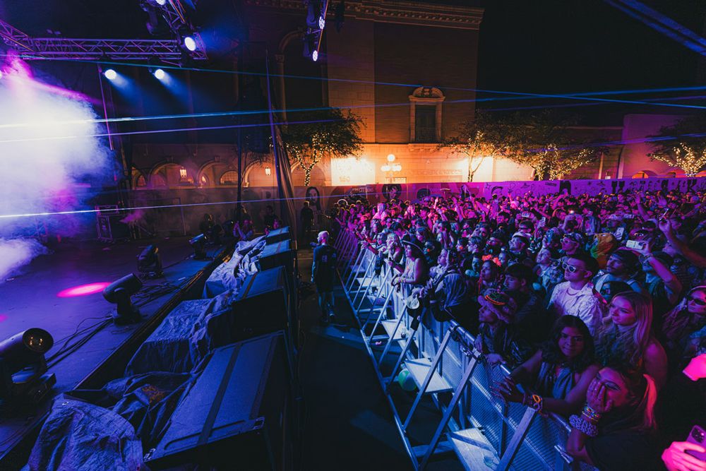 Large crowd at an indoor concert venue bathed in purple and pink lighting with stage equipment visible.