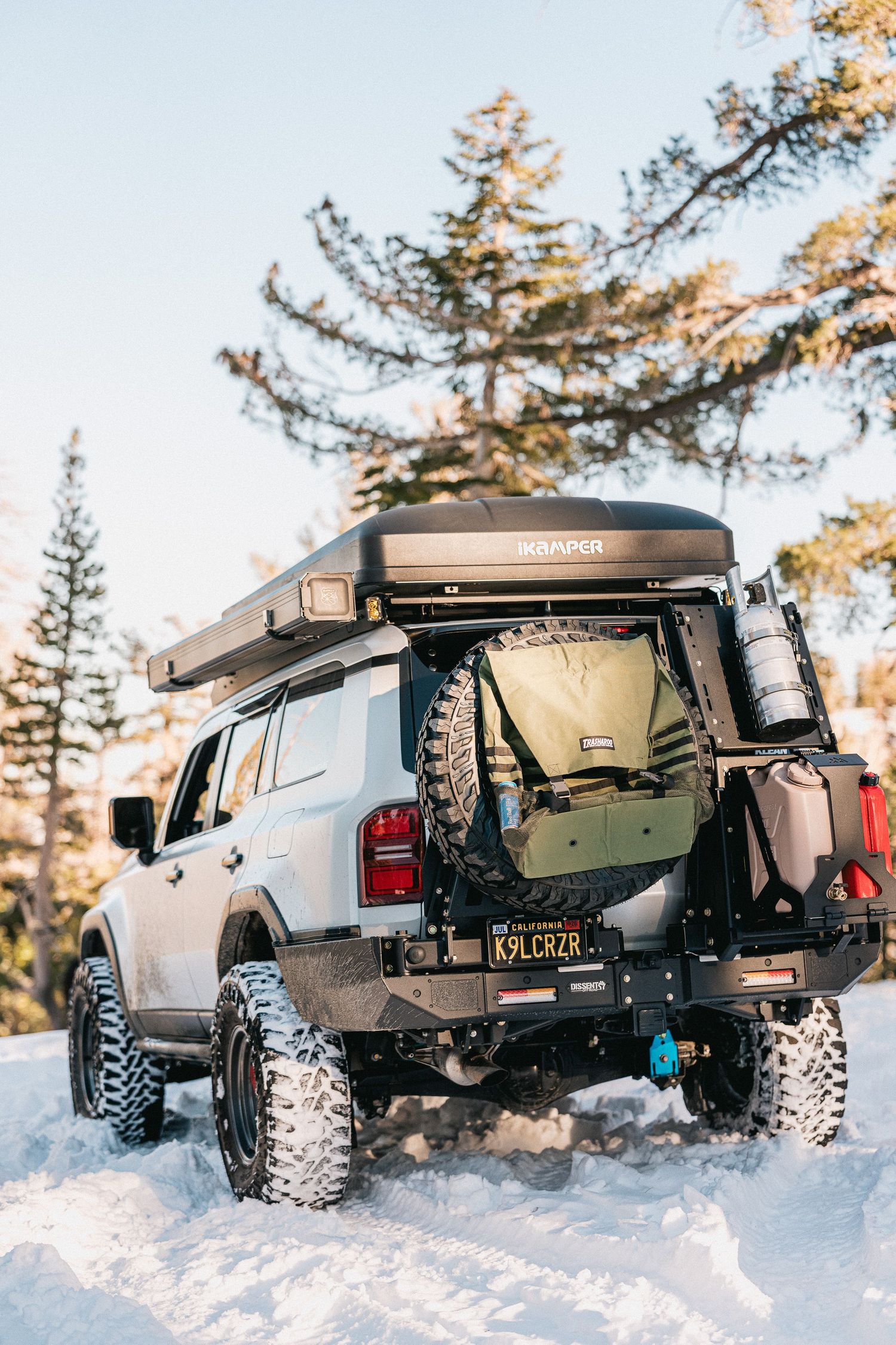 Black off-road truck driving through deep snow with large rugged tires against a backdrop of evergreen trees.