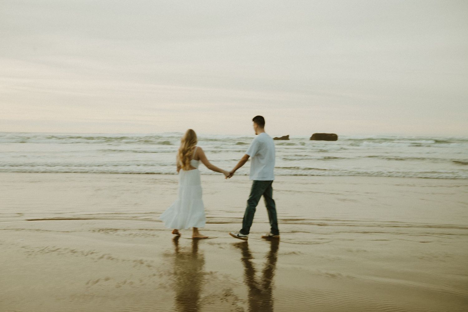Couple walking along a serene beach at sunset with rocky cliffs in the distance.
