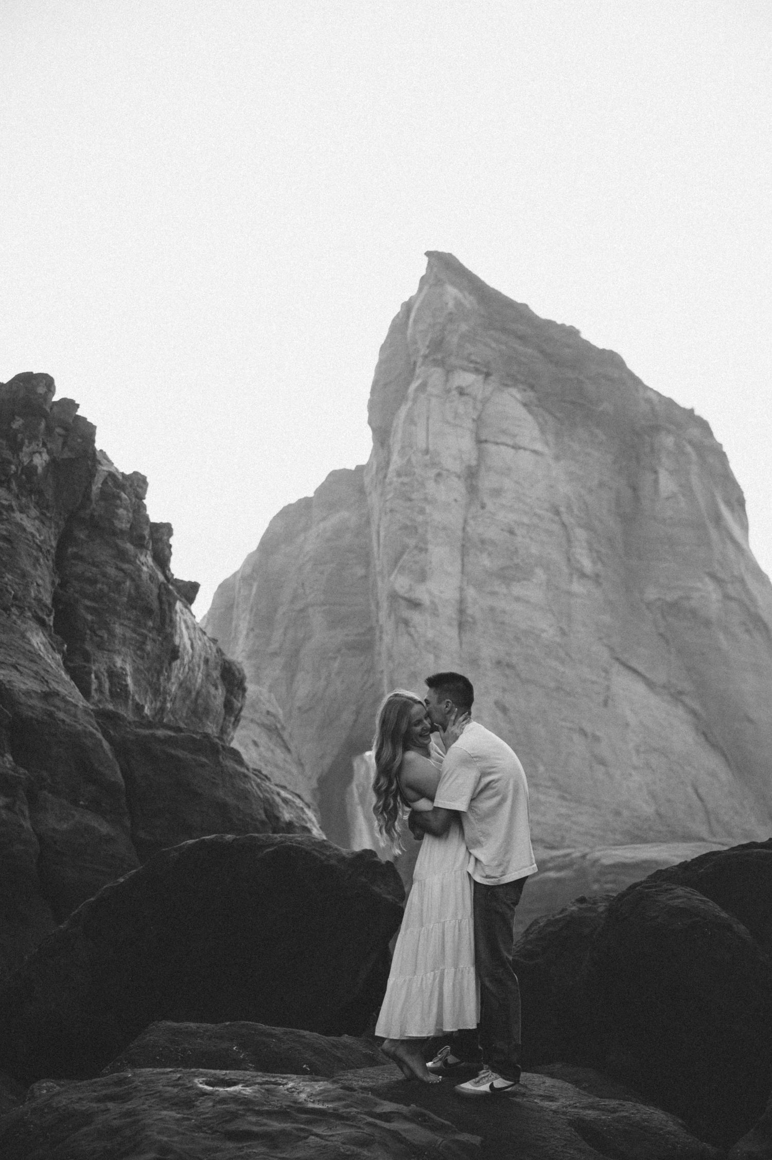 Black and white photo of a couple standing on dramatic rock formations with towering cliffs behind them.