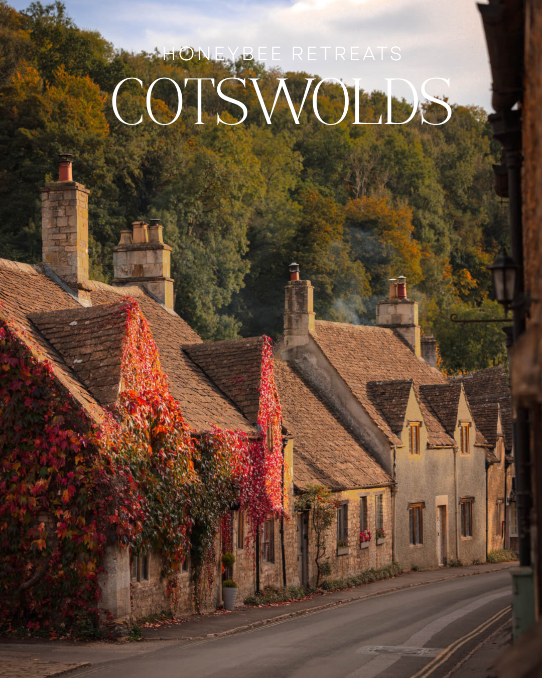 Charming Cotswolds stone cottages draped in vibrant autumn ivy lining a quiet village street with lush green hills behind.