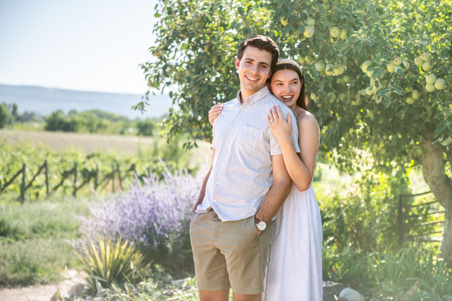A newly engaged young couple embracing outdoors near vineyard and lavender, smiling in summer sunshine.