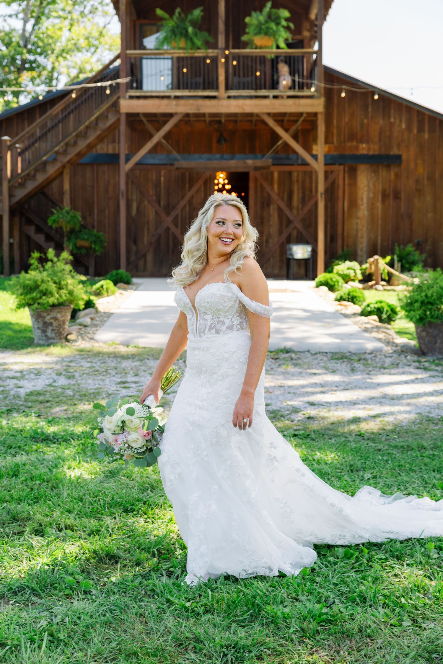 A wedding dress flows gracefully across green grass near a rustic wooden barn on a sunny summer day.