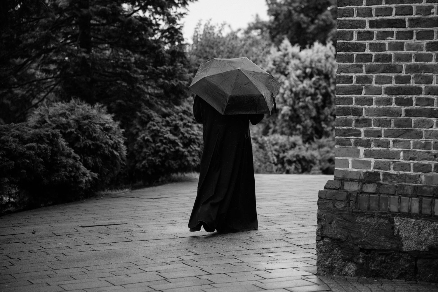 A figure in a dark cloak walks along a stone path next to a brick wall in a black and white photograph.