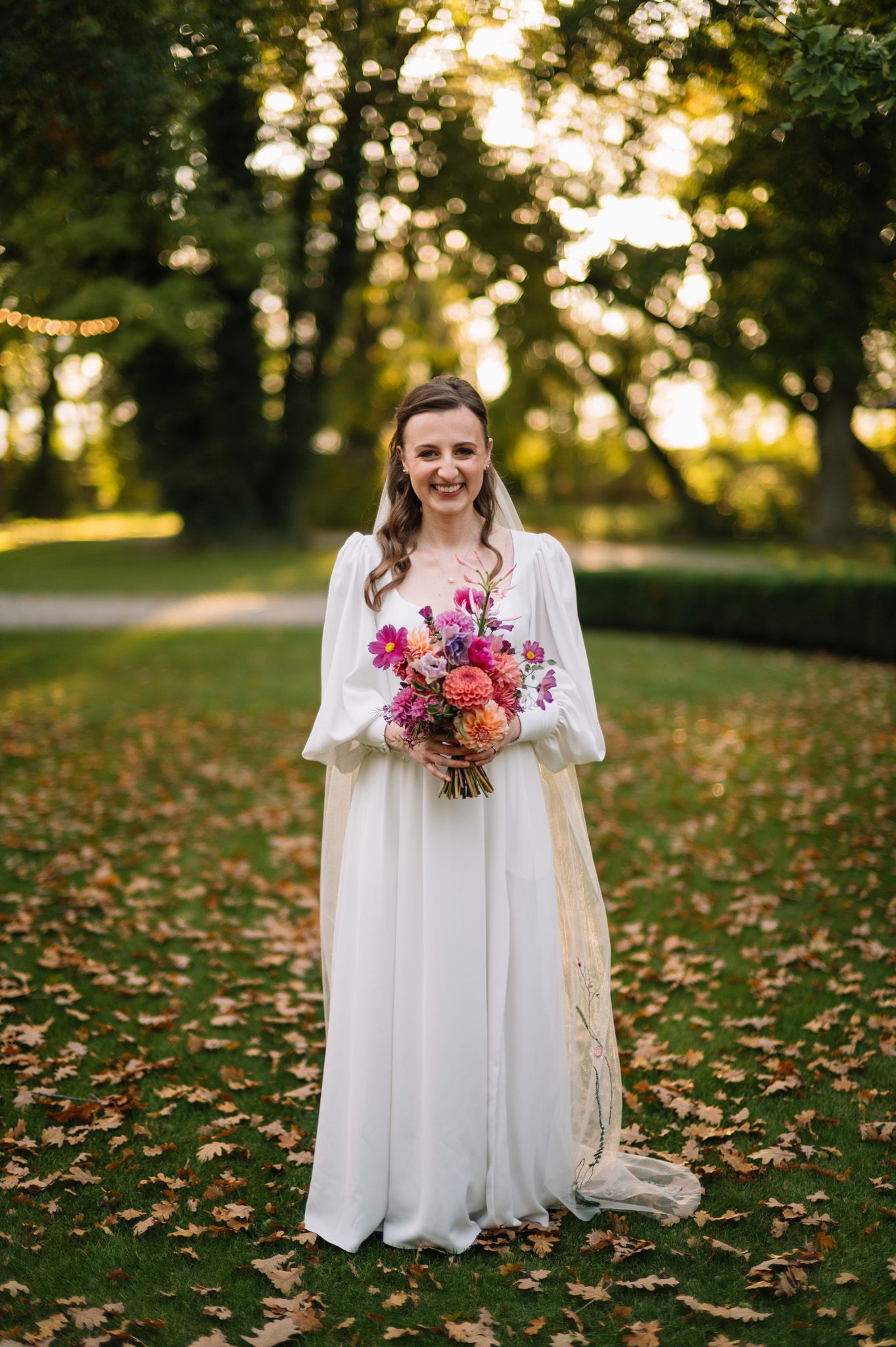 A bride in a flowing white dress holds a vibrant pink and coral bouquet while standing on autumn leaves at sunset.