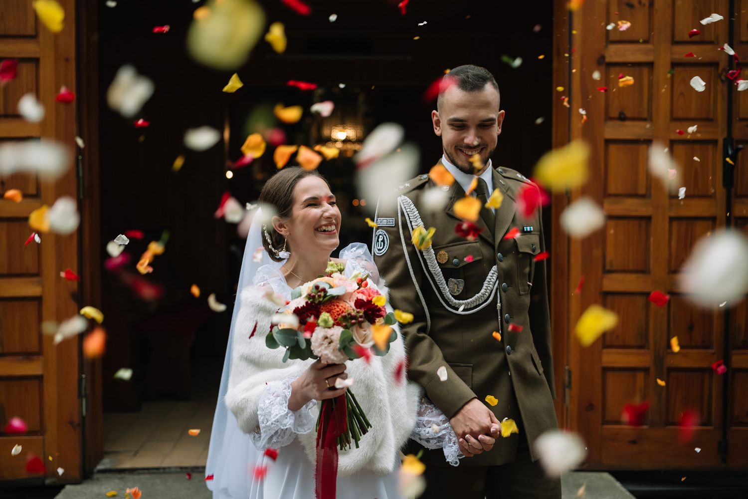 Wedding guests throw colorful confetti over a couple exiting through wooden church doors in celebration.