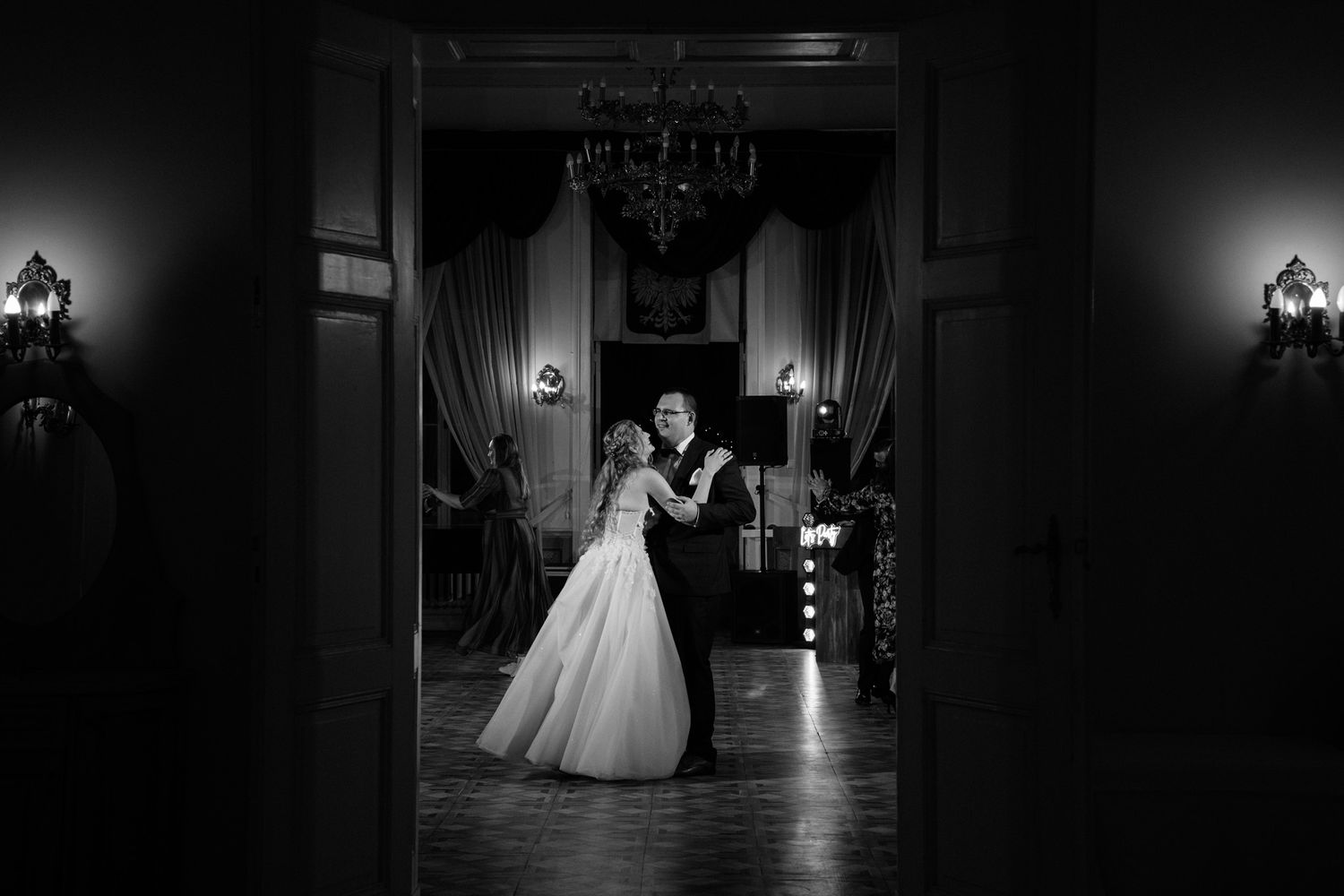 Black and white wedding photograph showing a bride in a ballgown silhouetted against dramatic lighting in an elegant venue.