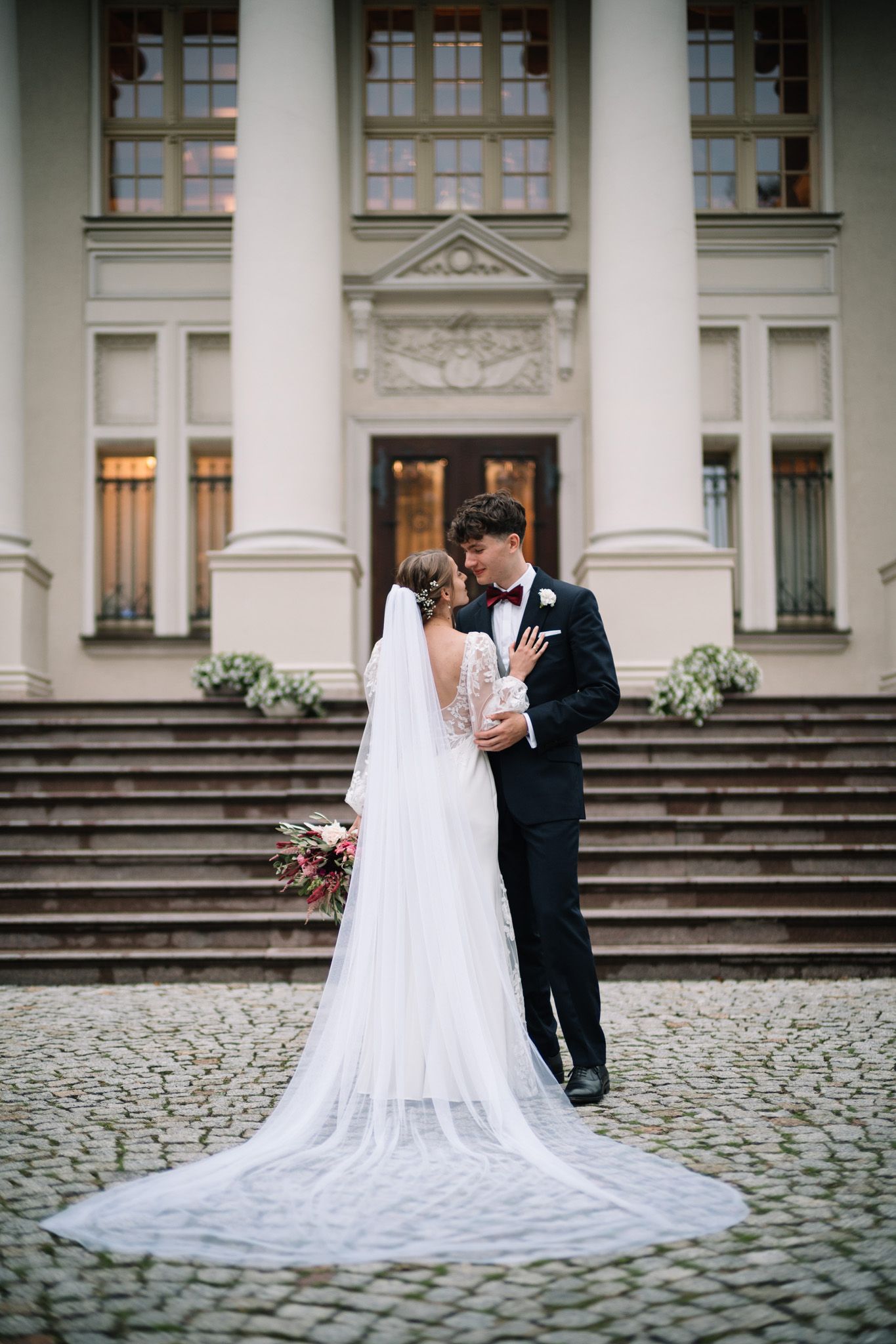 A romantic wedding couple stands on cobblestone steps in front of a grand building with white columns and a flowing veil.
