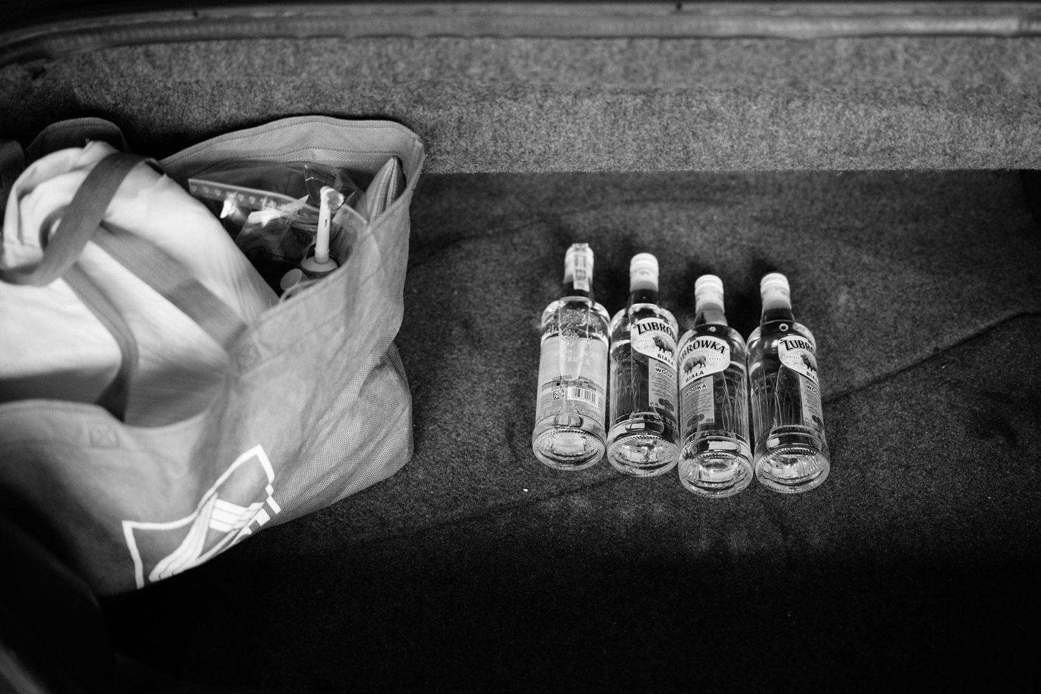 Black and white photo of empty glass bottles lined up next to a white plastic bag on a dark surface.