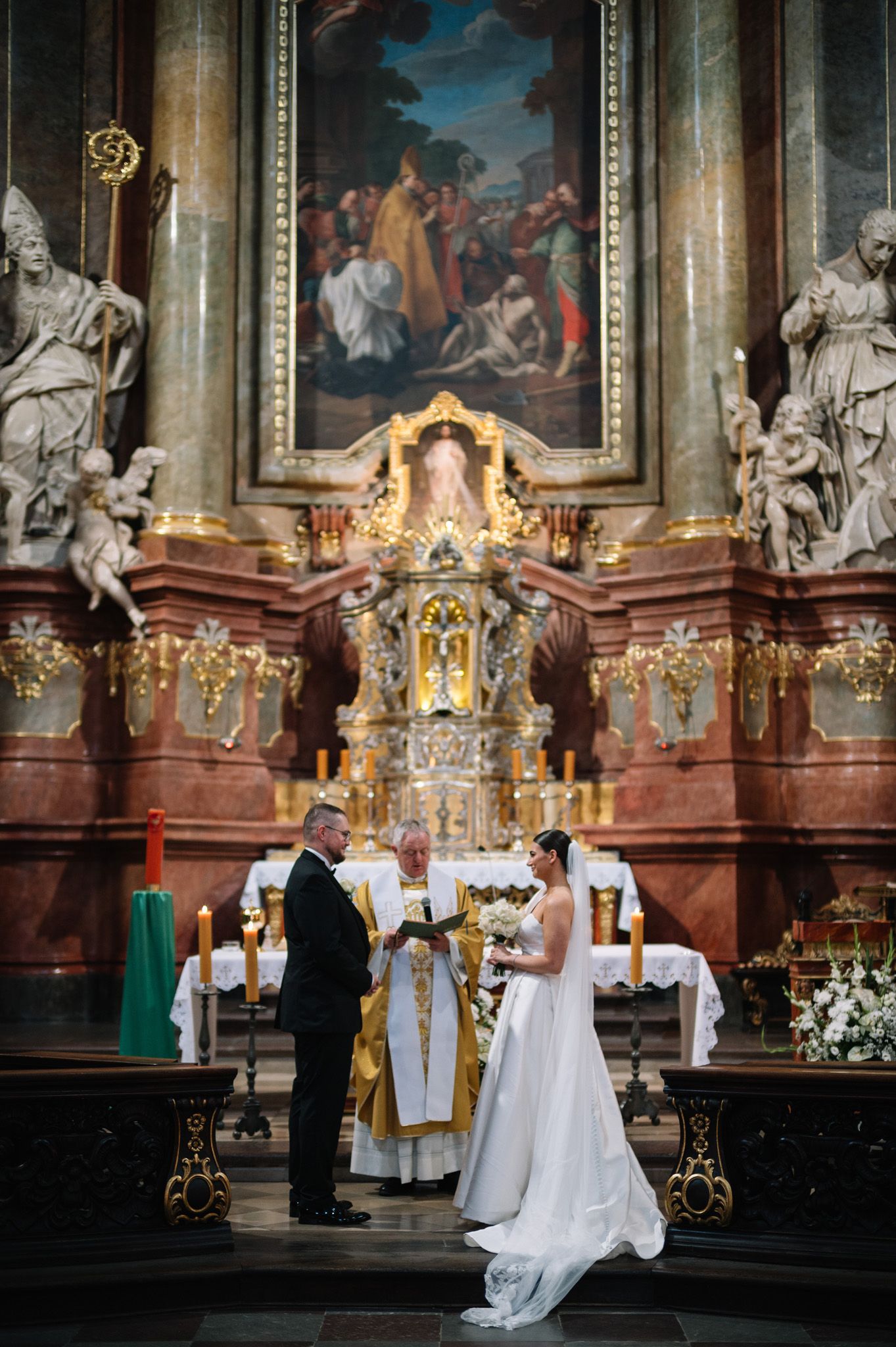 A wedding ceremony takes place in an ornate Catholic church with baroque decorations, religious paintings and golden details.