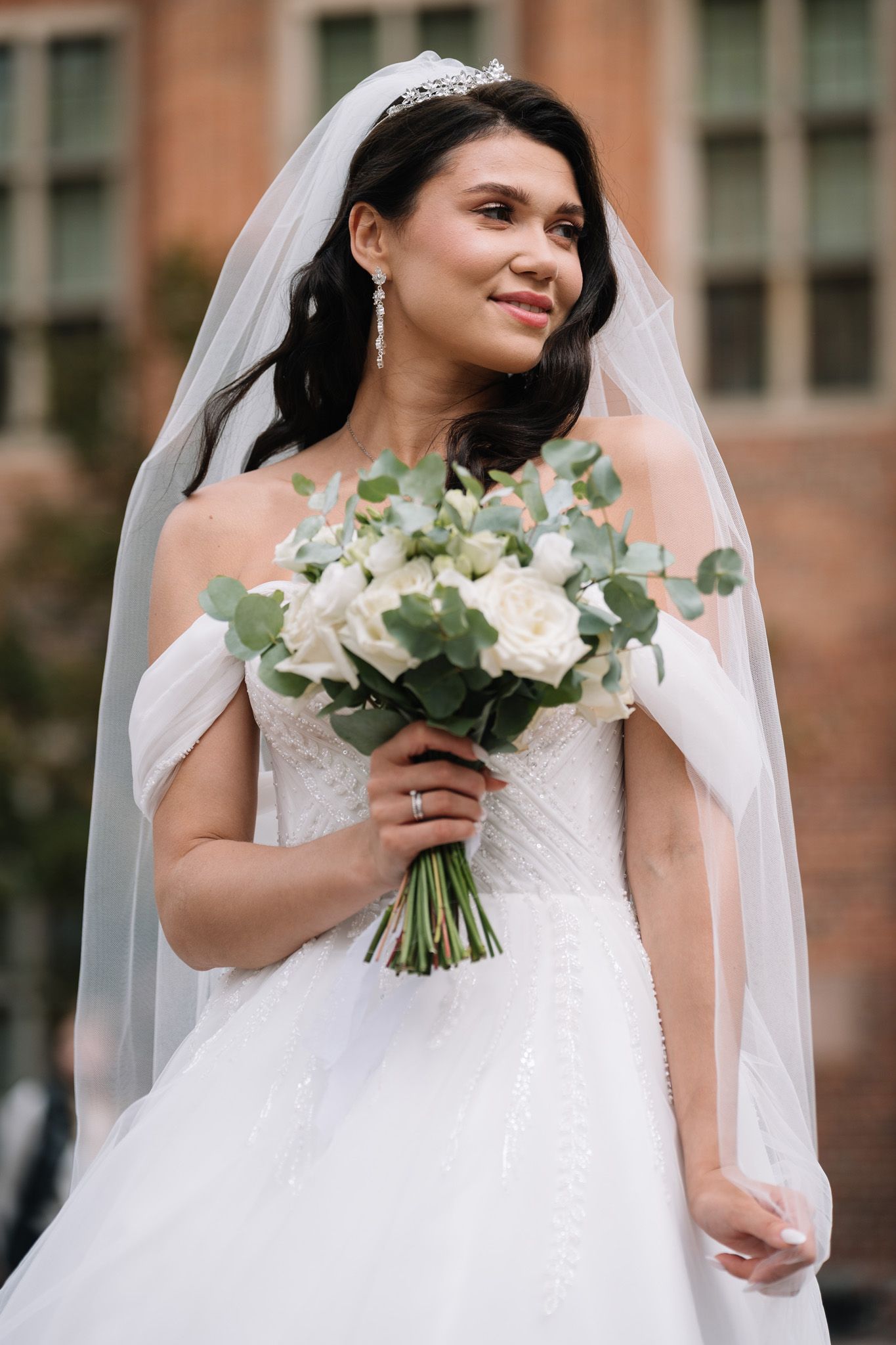Beautiful bride in white wedding dress holds elegant bouquet of white flowers and eucalyptus against brick building.