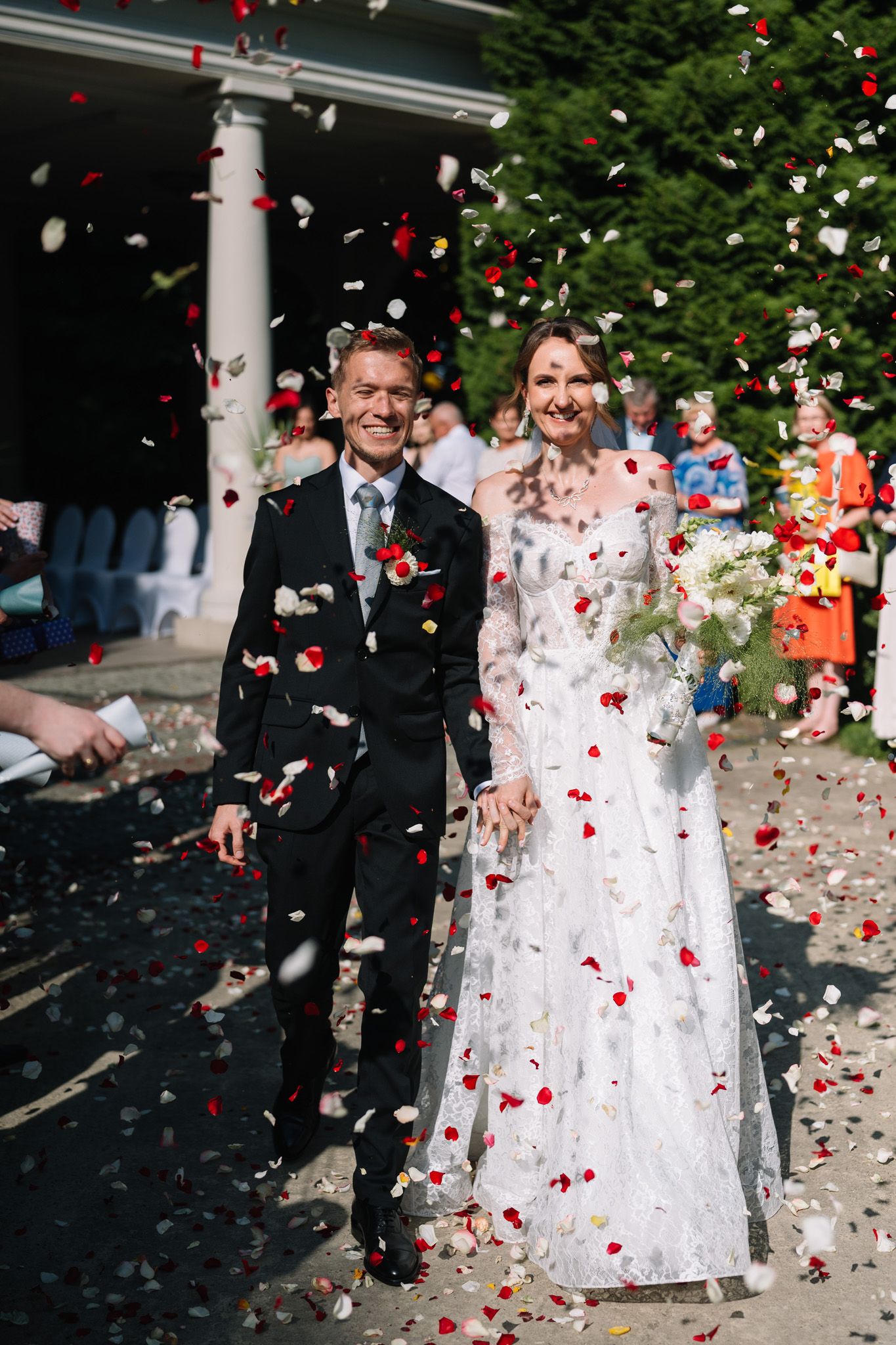 A joyful wedding couple stands under white columns as red and white confetti rains down around them.