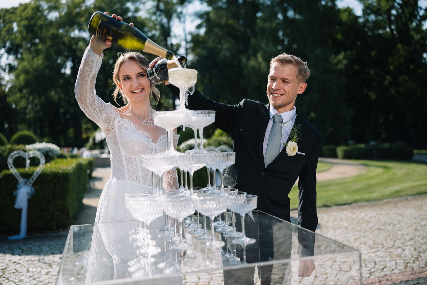 A couple pours champagne into a tower of glasses during an elegant outdoor wedding celebration.