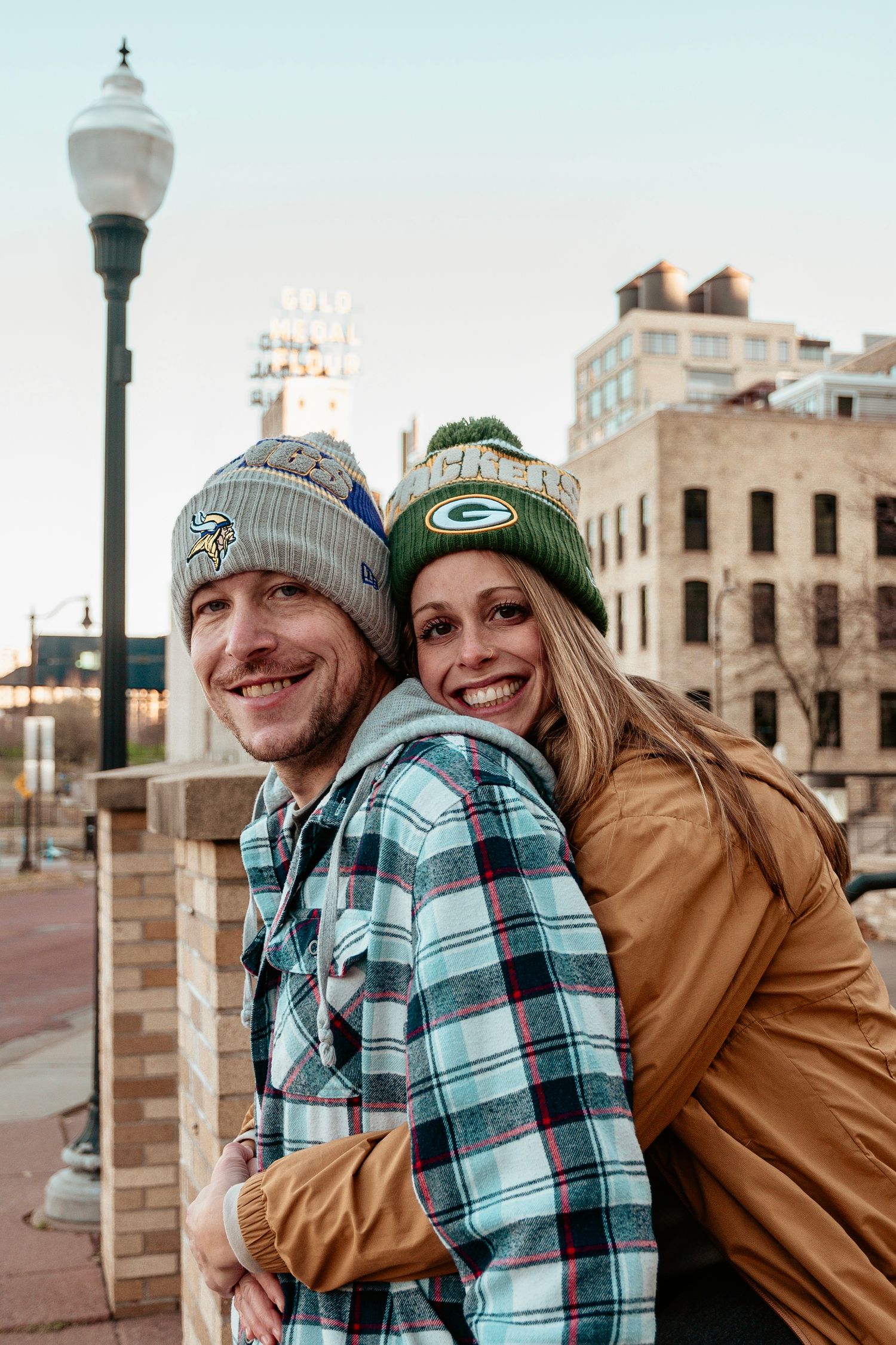 A couple wearing winter hats and plaid clothing share playful moments together outdoors in an urban setting.