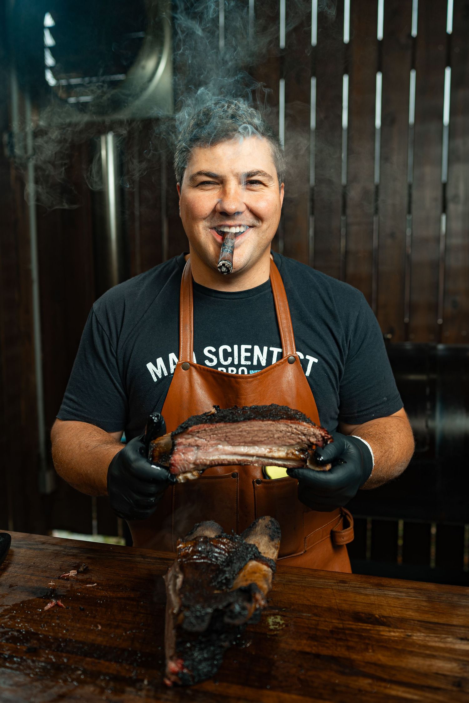 BBQ chef in black t-shirt and apron shows off smoked beef ribs on wooden cutting board.