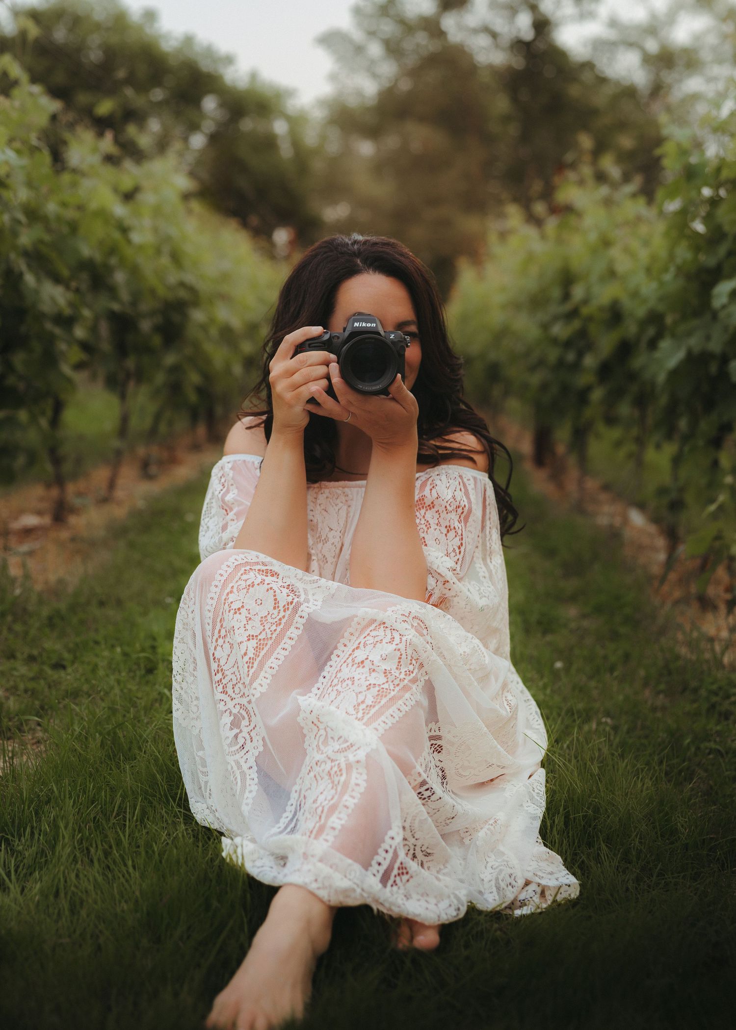 Photographer in white lace dress taking photos in vineyard setting.