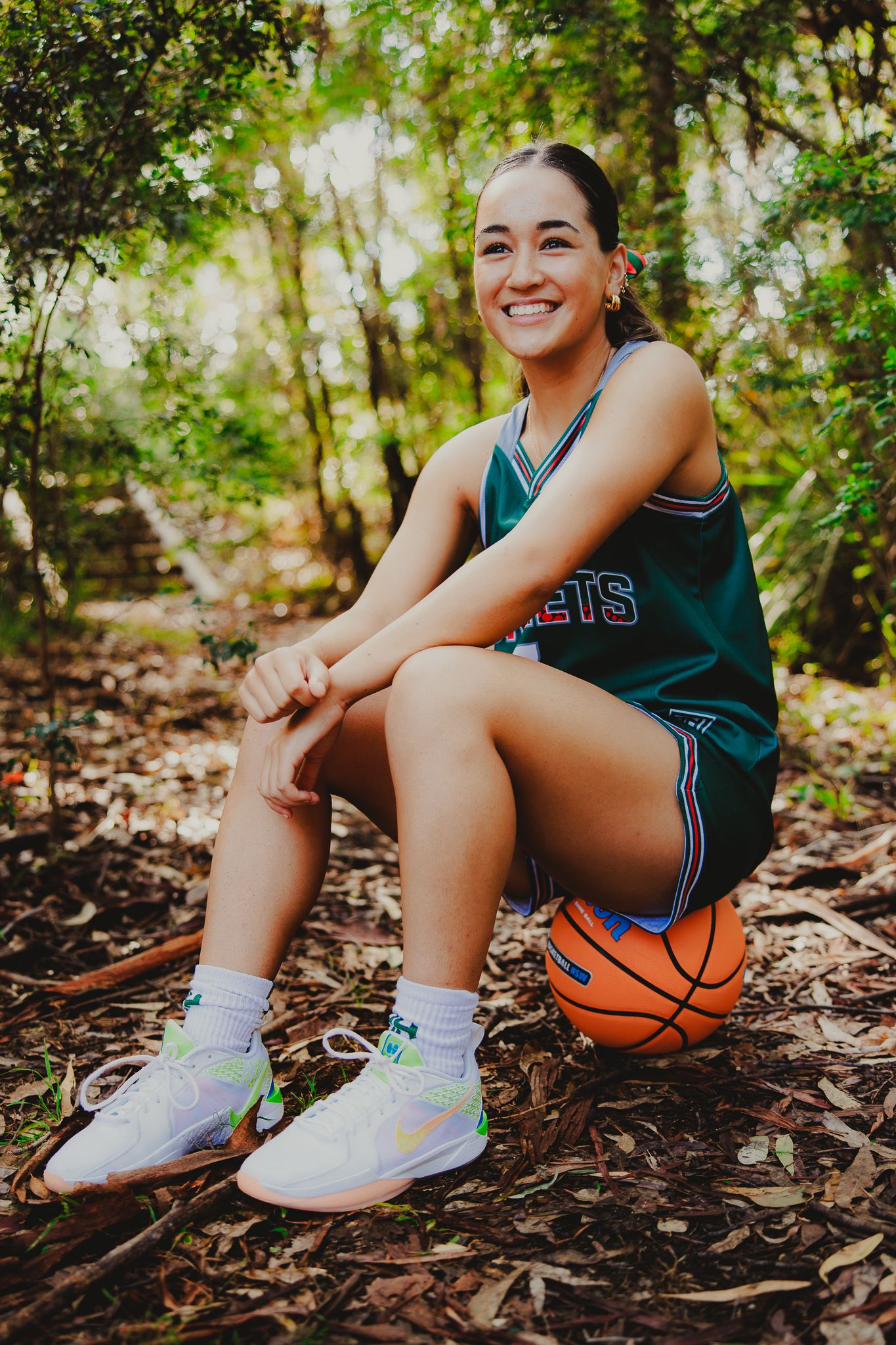 Basketball player sits on forest ground with basketball and colorful sneakers.