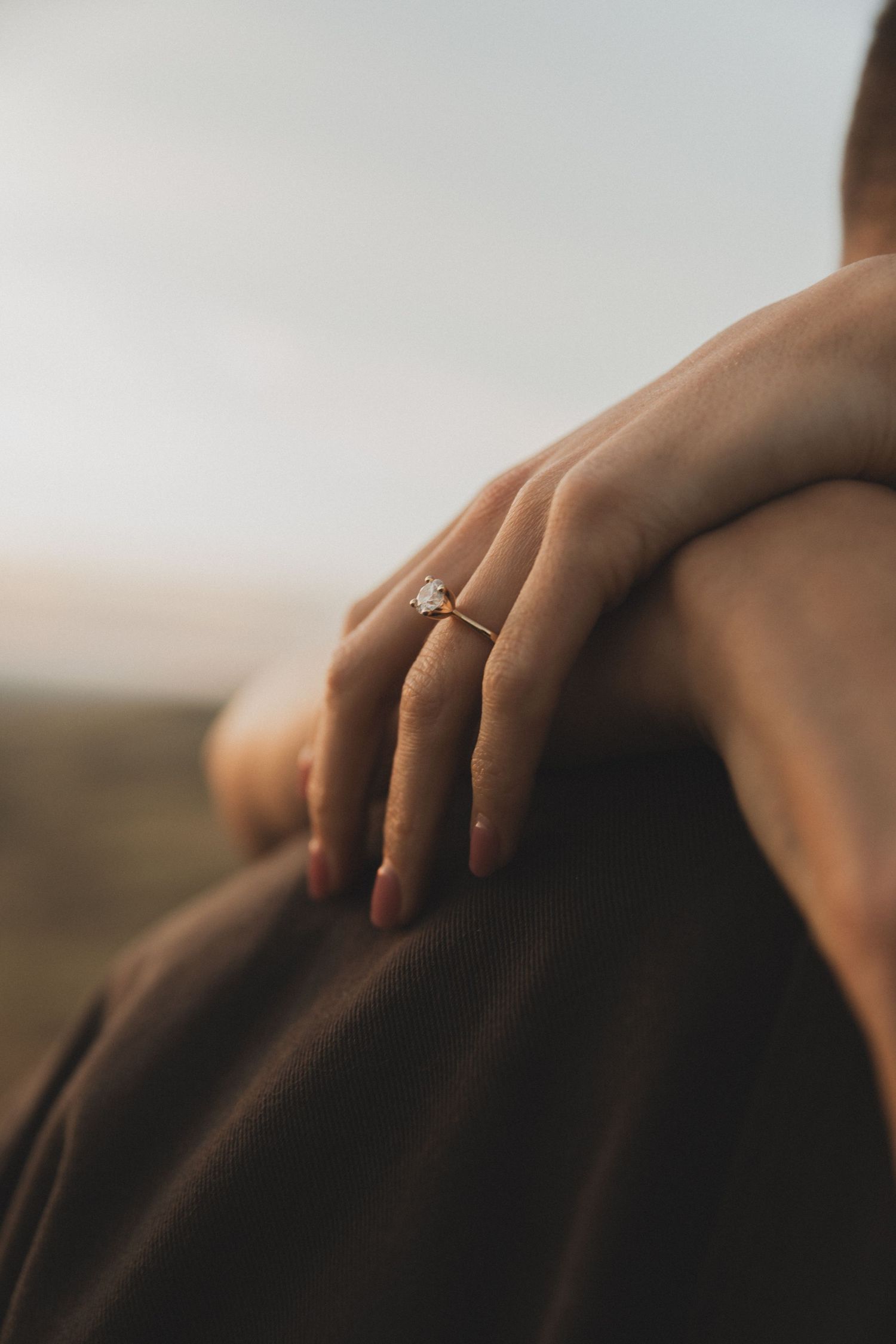 A close-up view of hands wearing an engagement ring resting gently on a dark surface in warm lighting.