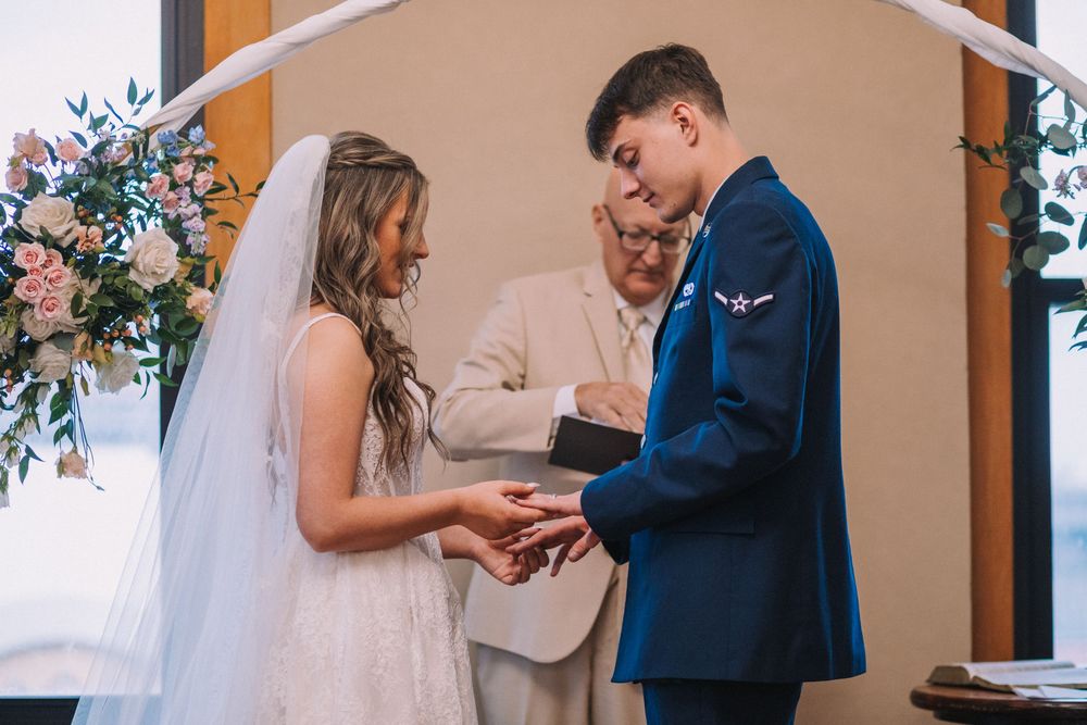 Bride and groom exchange rings during wedding ceremony with floral arrangements.