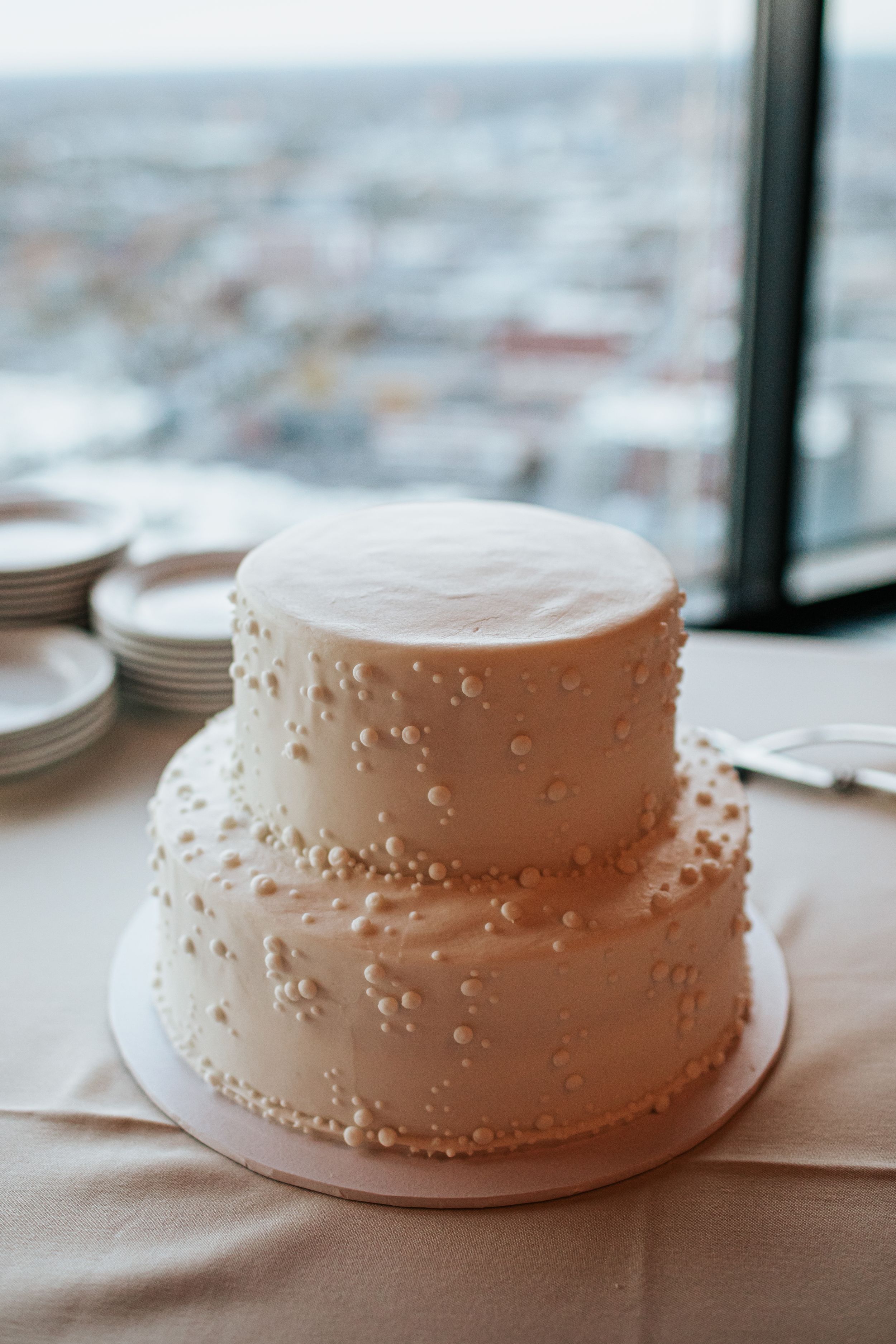 A white frosted cake with delicate piped details sits on a wooden surface near a window with natural lighting.