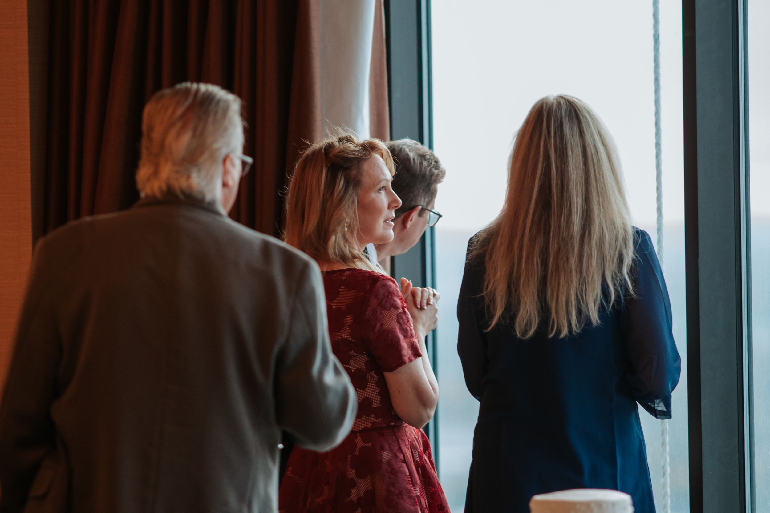 People gather near large windows in an office or meeting space, creating silhouettes against natural lighting.