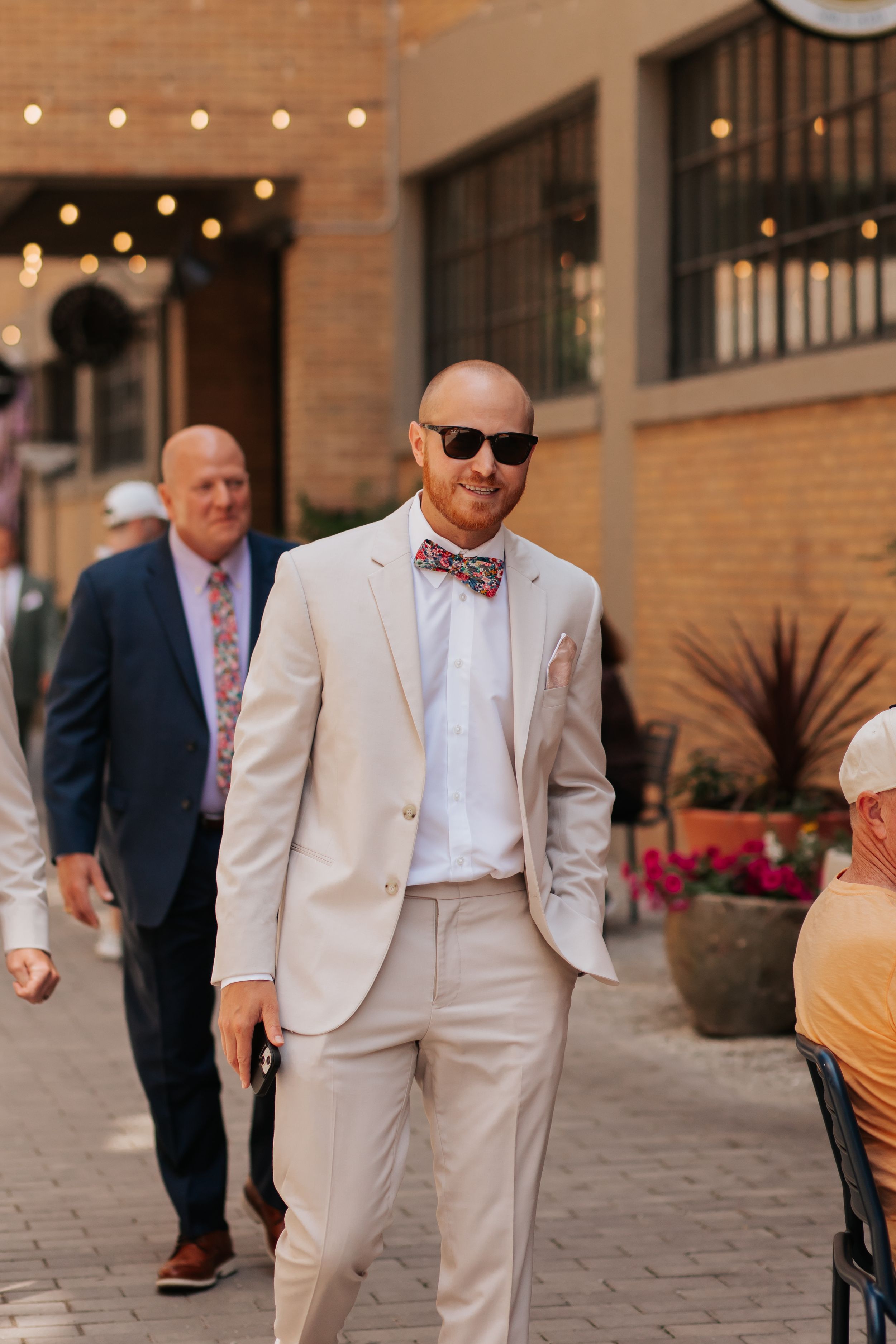 Well-dressed man in beige suit and bow tie walking outdoors with string lights and brick building in background.