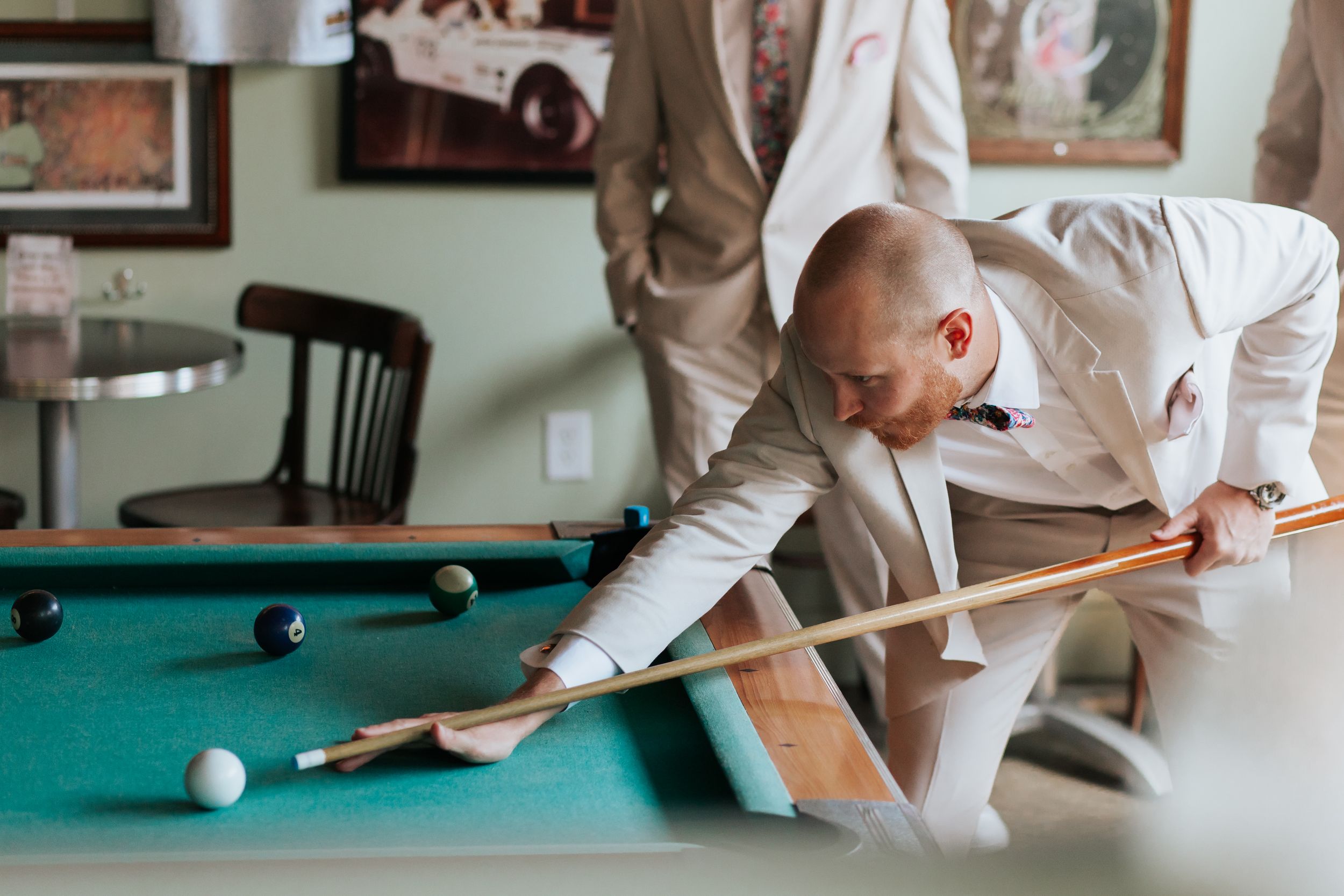 A person in a light colored suit leans over a pool table to take a shot during a game of billiards.