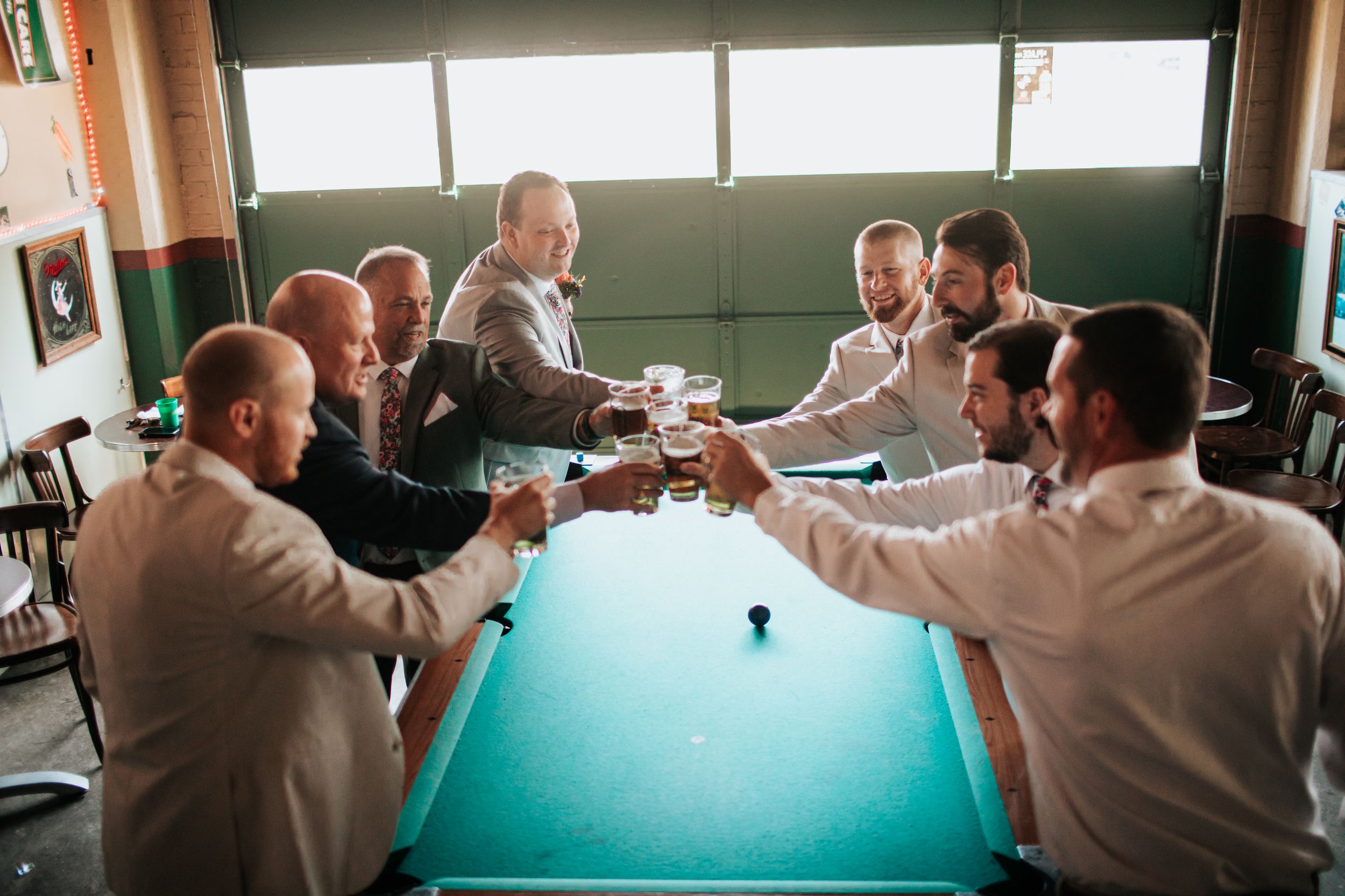 People in formal attire gather around a pool table during what appears to be a wedding celebration.