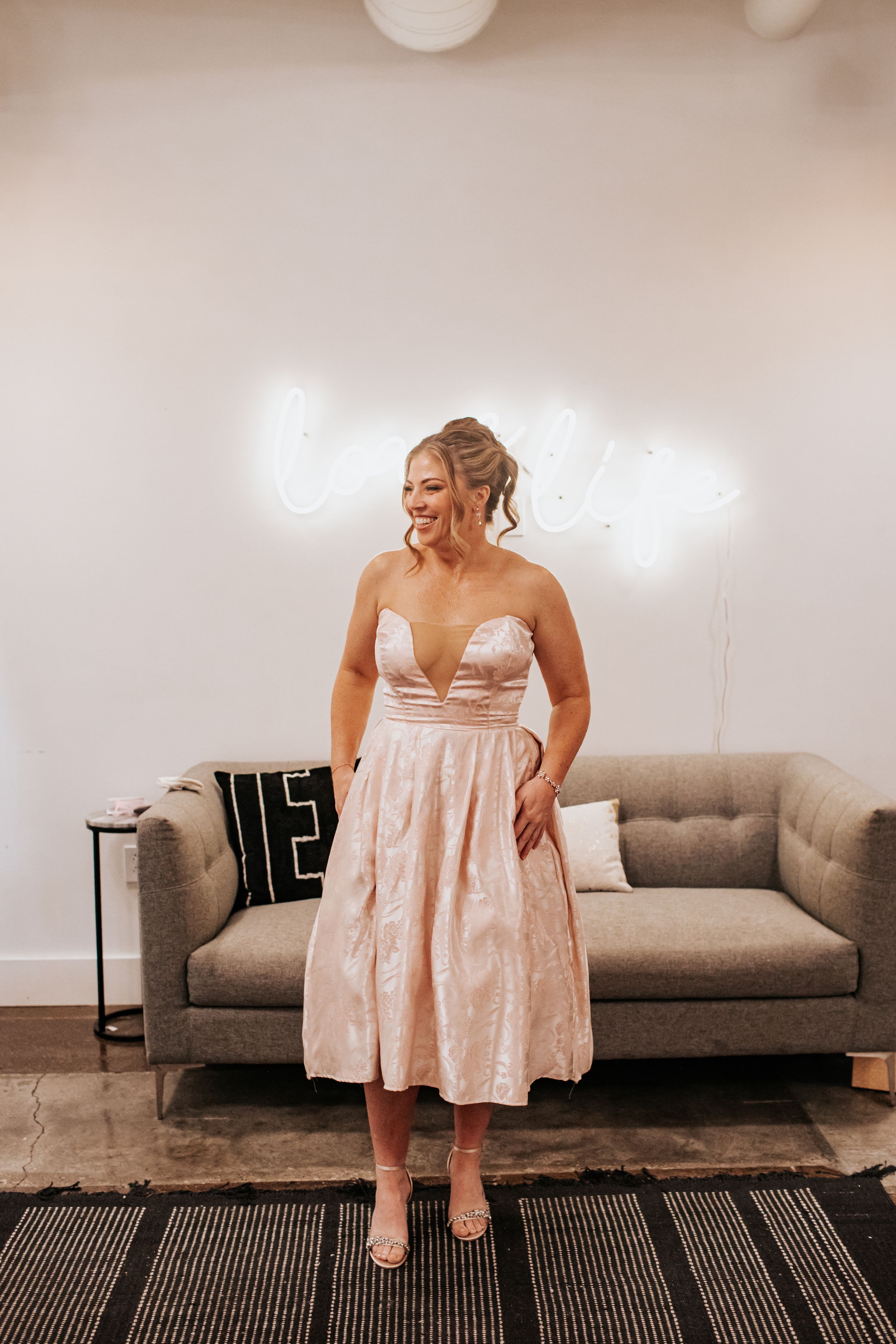 Person in a blush pink dress sitting and posing playfully on a grey couch.