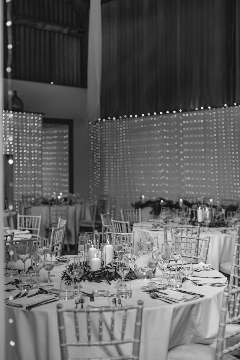 Black and white photo of wedding reception tables with fairy light backdrop and elegant place settings.