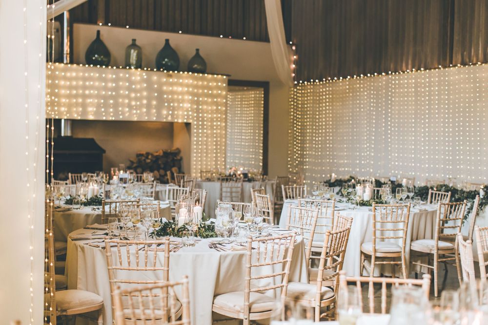 Wedding reception venue features white chairs and elegant table settings with crystal chandeliers overhead.