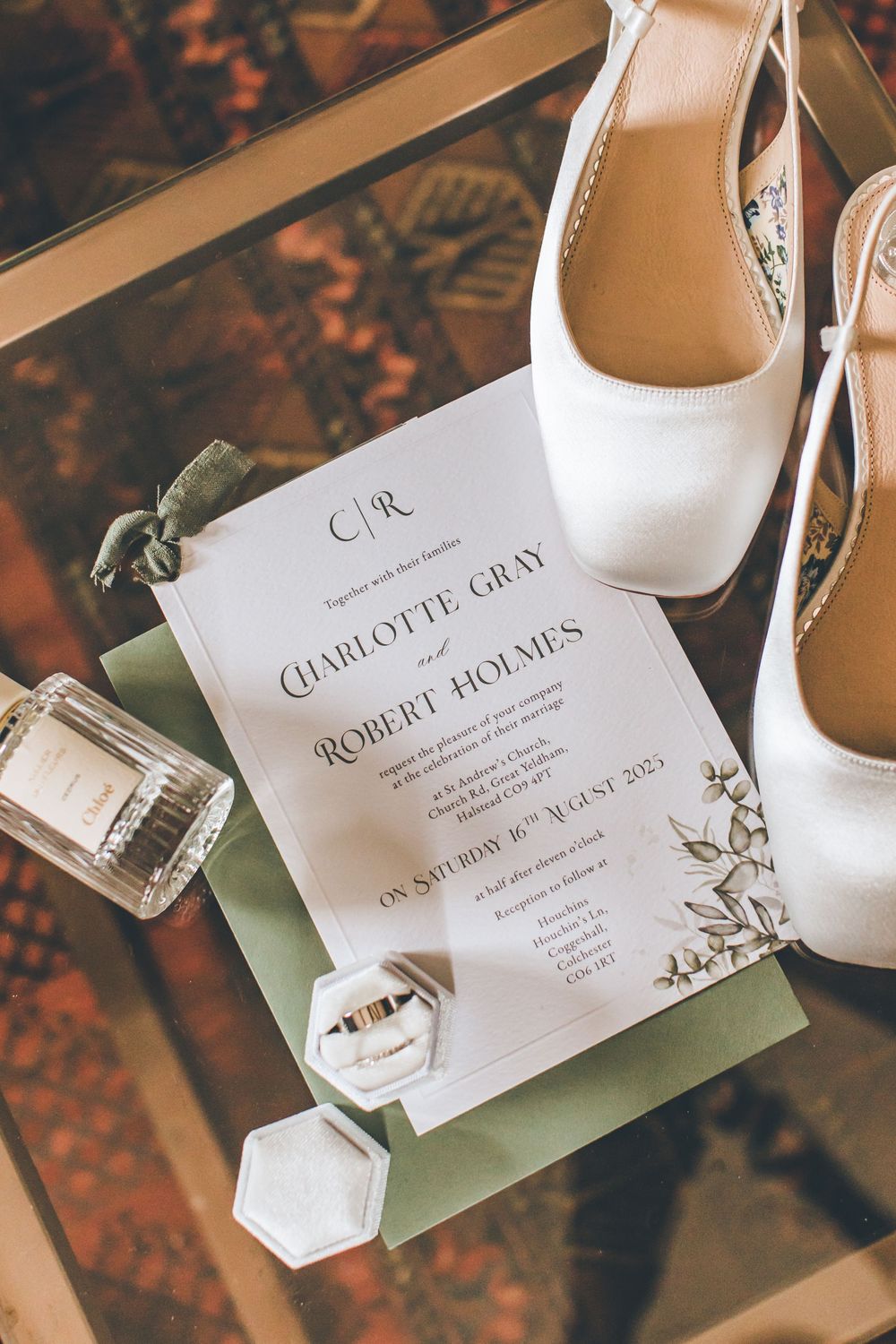 Wedding invitation with white bridal shoes, perfume bottle and green accents on a patterned surface.