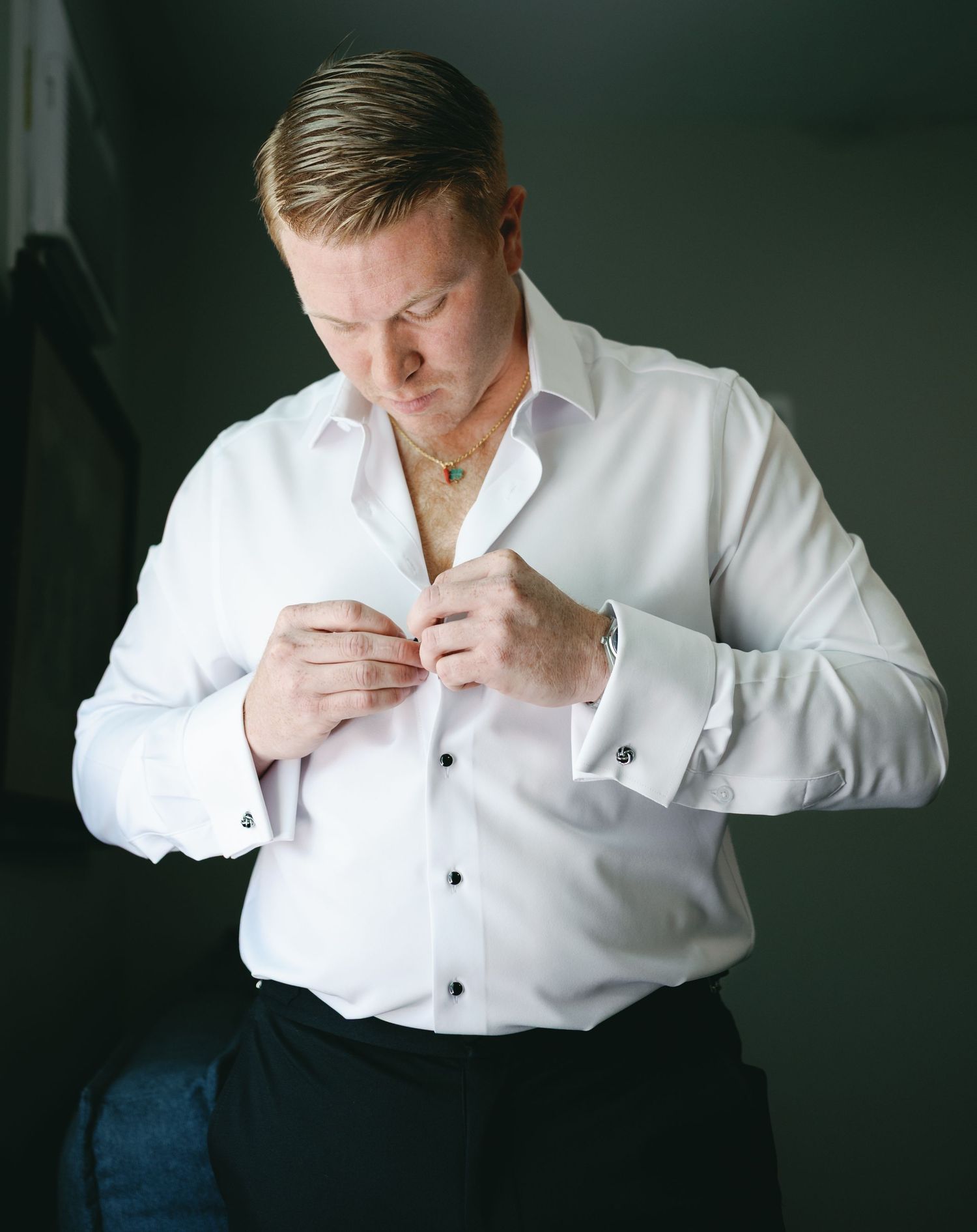 Someone buttoning up a crisp white dress shirt while wearing a gold necklace against a dark background.