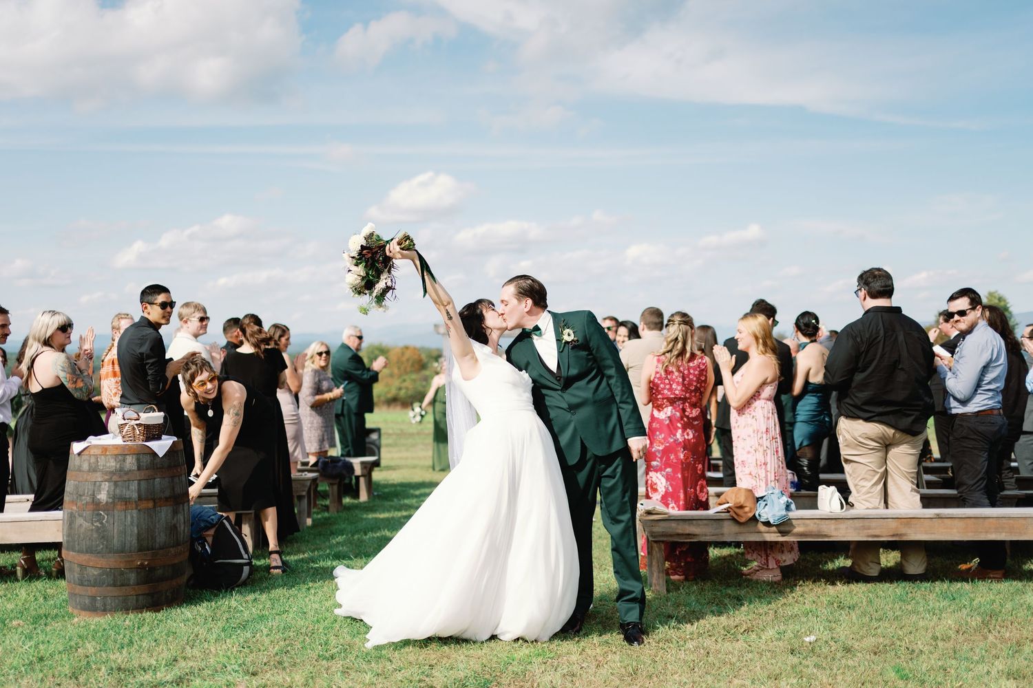 Outdoor vineyard wedding celebration with guests gathered around wine barrels on a sunny day.